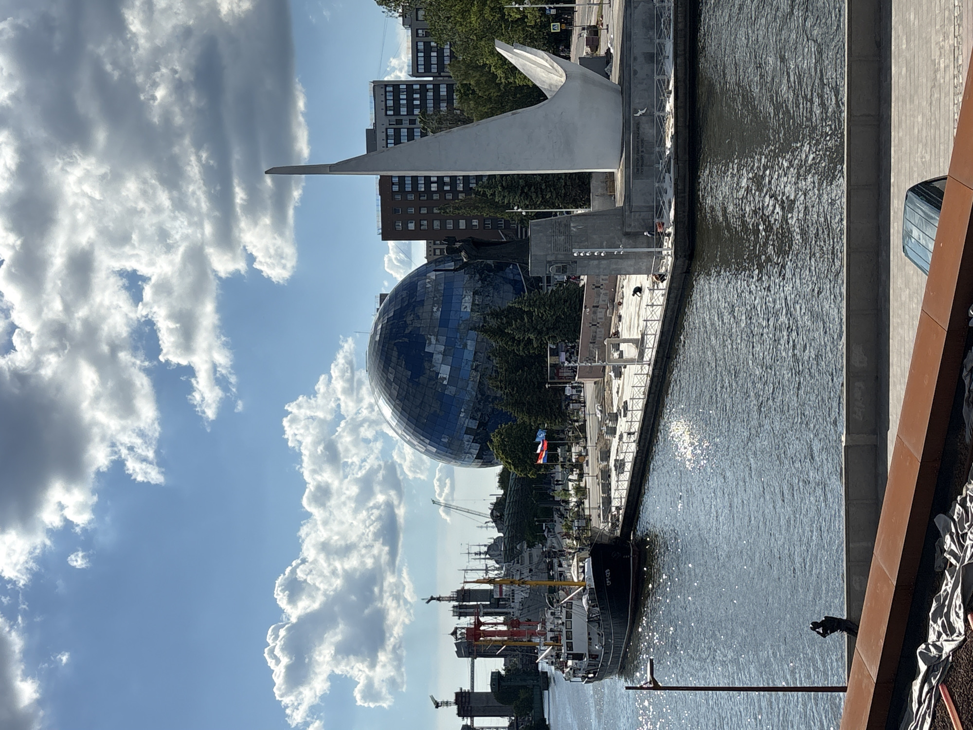 This image presents a sunny daytime scene of a waterfront in Kaliningrad, Russia, featuring a combination of urban architecture, maritime elements, and striking landmarks.
In the midground, a prominent feature is a large, iridescent blue spherical building, likely the main building of the Museum of the World Ocean, its surface reflecting the bright blue sky and fluffy white cumulus clouds. Adjacent to it, a tall, abstract white monument with a soaring, pointed form, reminiscent of a ship's prow or a stylized wave, stands on a concrete base. Faint Cyrillic text can be observed on the monument's pedestal. A Russian flag, with its white, blue, and red horizontal stripes, flies nearby.
The right side of the frame is dominated by a body of water, which shimmers intensely under the direct sunlight, displaying ripples across its surface. Along the water's edge, a paved embankment or promenade hosts several docked boats and ships. One vessel in particular clearly displays the identifier "ОПЧ-623" on its side. In the distance, further along the waterway, additional boats and industrial cranes suggest a bustling port area.
The background reveals a cityscape with various multi-story buildings, including residential blocks and commercial structures, interspersed with green trees. The sky is bright blue with scattered large, white cumulus clouds, indicating clear and pleasant weather. A few indistinct figures of people are visible on the promenade, suggesting normal pedestrian activity. In the immediate foreground on the far right, a small, dark, abstract sculpture of a human-like figure appears to be gazing out over the water. The scene conveys a sense of vibrant urban life intertwined with maritime heritage under excellent weather conditions.