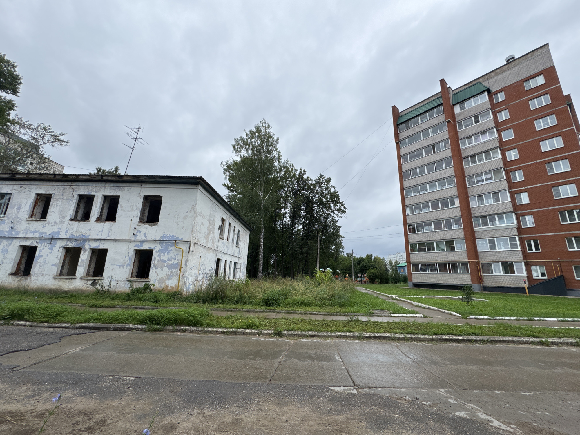The image shows a street scene in Chaikovsky, Russia, on an overcast day. To the left is a dilapidated, two-story building with peeling white paint and empty window frames, suggesting it is abandoned or under renovation. The building has graffiti on its exterior. Adjacent to this building and behind it are trees. To the right of the abandoned building is a modern, multi-story brick apartment building with numerous windows. Between the two buildings, there is a grassy area with some weeds and trees. A paved sidewalk and road are visible in the foreground, with wet patches indicating recent rain. In the distance, behind the apartment building, a playground and other residential buildings are partially visible.