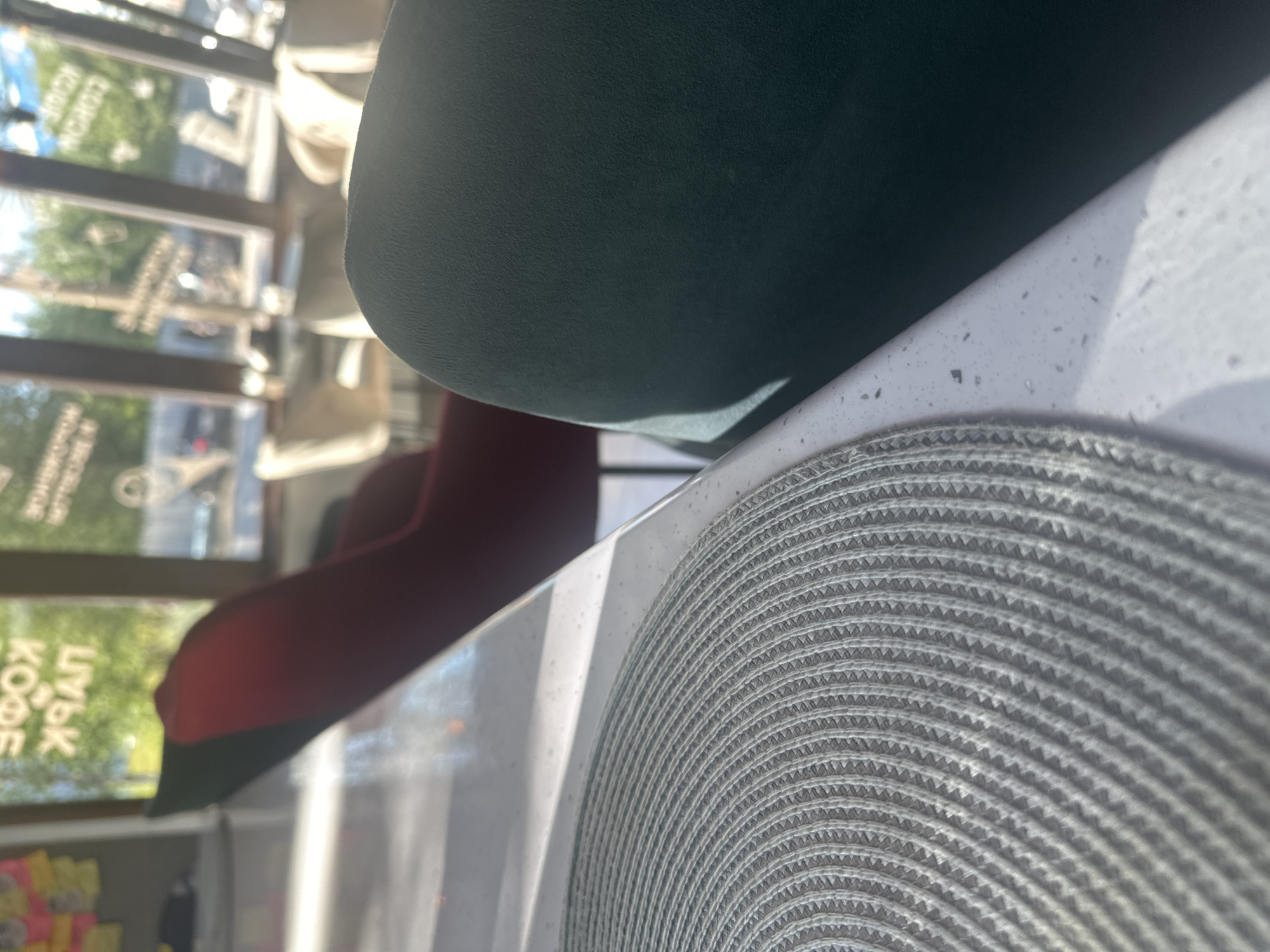 An indoor shot focuses on a white table with a gray circular placemat in the foreground. To the right of the placemat, a dark green upholstered chair is visible. In the background, through a large window, a red chair is partially visible. Outside the window, trees and buildings can be seen, along with a sign that appears to read "КОФЕ ПАРК" (Coffee Park) in Russian, indicating the location is likely a cafe or restaurant in Plesetsk, Russia. The lighting suggests it is daytime.