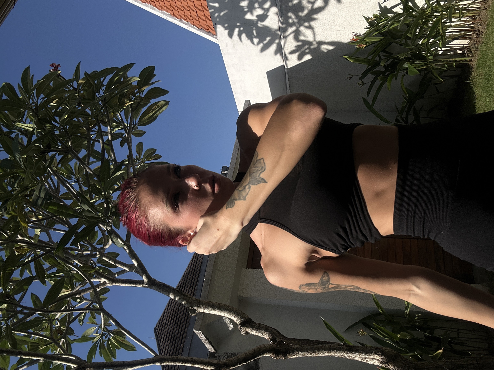 A medium-shot depicts a woman in athletic wear, seemingly posing outdoors. The woman has reddish-pink hair and tattoos on her arms. She is wearing a black sports bra and matching bottoms. A tree with thick foliage fills the left side of the frame, while a building with a white facade and red roof tiles is visible in the background. Additionally, a green shrub and a patch of grass can be seen on the right. It appears to be a sunny day. The location is in North Kuta, Indonesia.