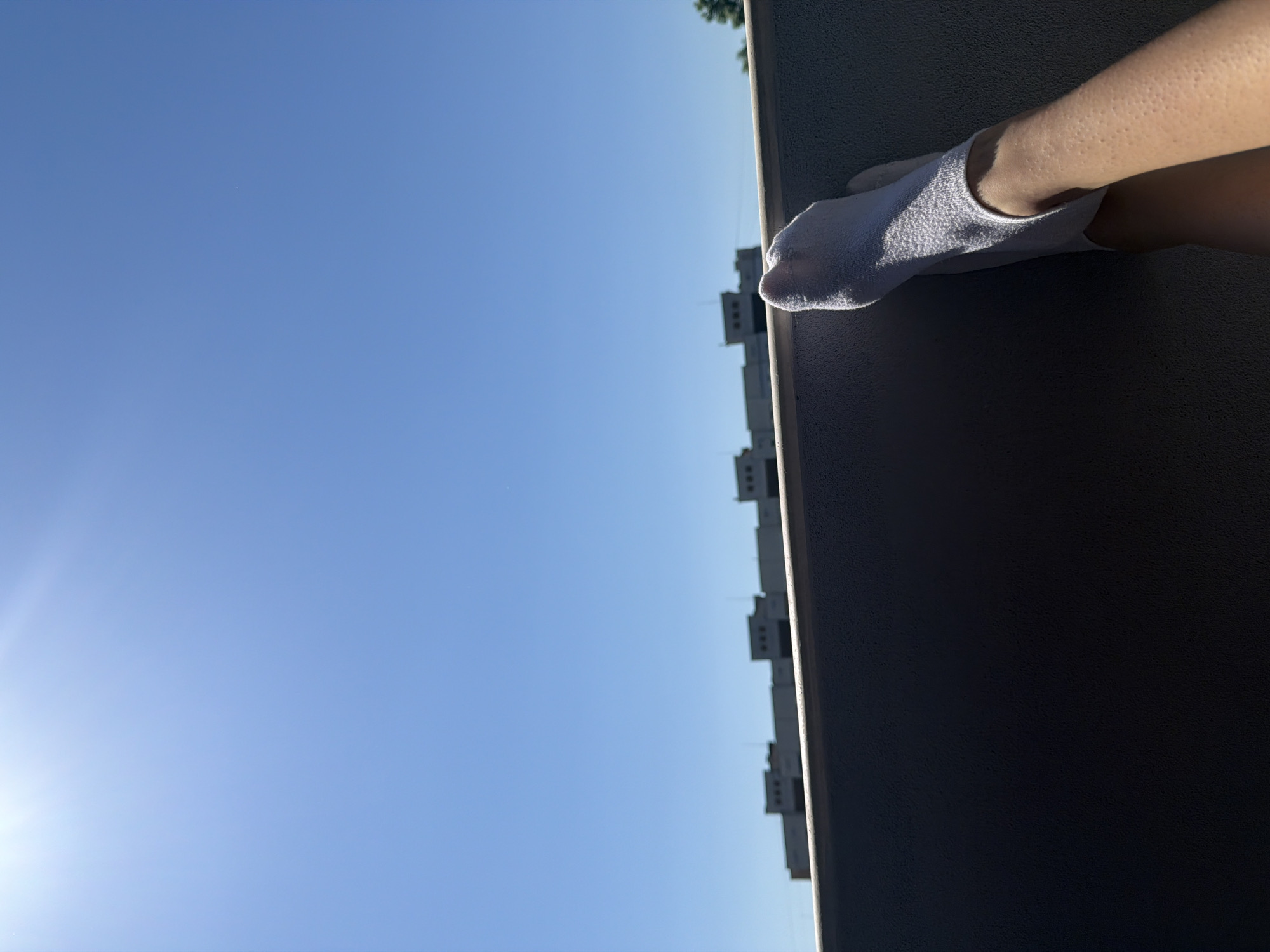 The image shows a close-up view of a person's legs and feet, wearing white socks, resting on what appears to be a balcony railing. The legs and feet are illuminated by sunlight, casting shadows on the surrounding dark surface. In the background, a clear blue sky stretches across the frame, with a line of buildings visible on the horizon. The architecture of the buildings suggests a setting in the city of Ruse, Bulgaria, characterized by their repetitive, modernist design. The overall scene evokes a sense of relaxation and leisure, likely taken outdoors during a sunny day.