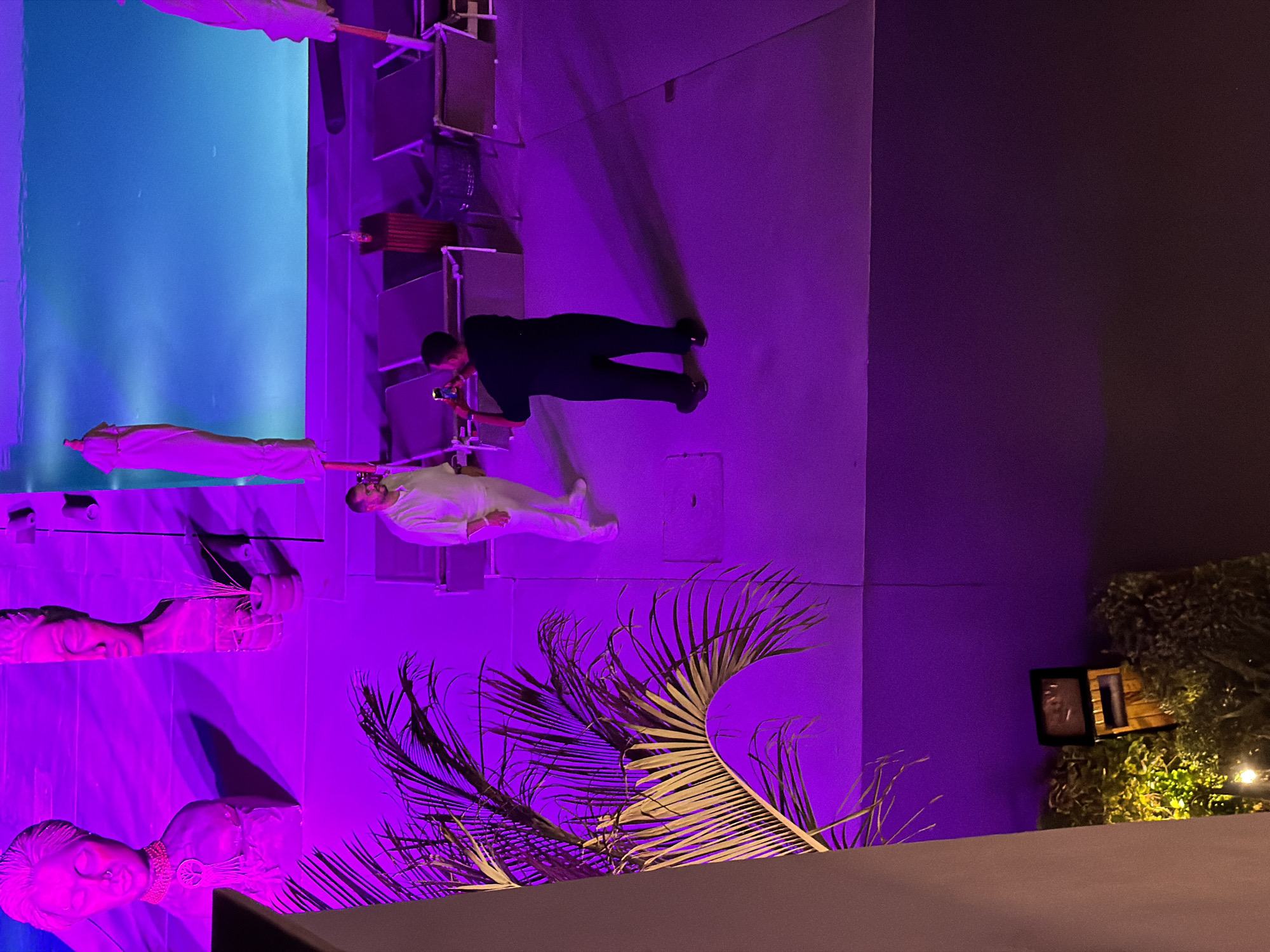 The image shows an outdoor scene at night, illuminated by purple lights. In the foreground, a person is standing and looking towards a pool area where another person is taking a picture with a phone. The pool is visible in the background with blue lighting. Lounge chairs and umbrellas are also present around the pool. To the left, a palm frond is prominent. The overall atmosphere suggests a resort or a similar leisure establishment. The city of Sharm El Sheikh, Egypt, is known for its resorts and beautiful pools, often illuminated with colored lights at night for aesthetic purposes.