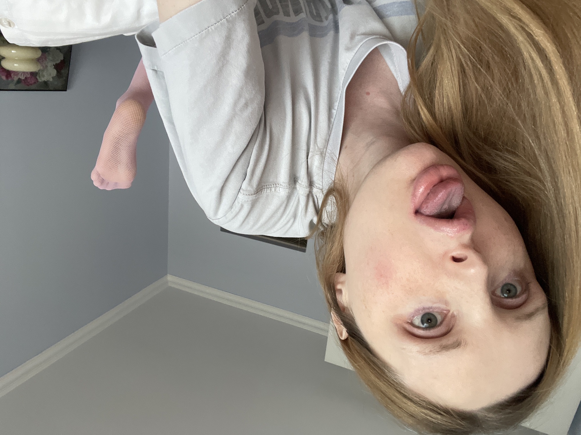 A close-up, low-angle, indoor, selfie-style shot shows a person with blonde hair lying down and sticking their tongue out. The person's face is the primary focus, with their eyes open and looking towards the camera. Their mouth is open, and their tongue is extended. Redness is visible on the cheeks. They are wearing a light-colored t-shirt. A pink fishnet stocking is visible on their right foot, which is slightly raised. The background consists of a light blue wall with white crown molding and a portion of a ceiling. A framed picture of flowers and decorative objects is visible on a surface in the lower right corner. The scene does not provide any specific details to indicate its location within Yablonovskiy, Russia.