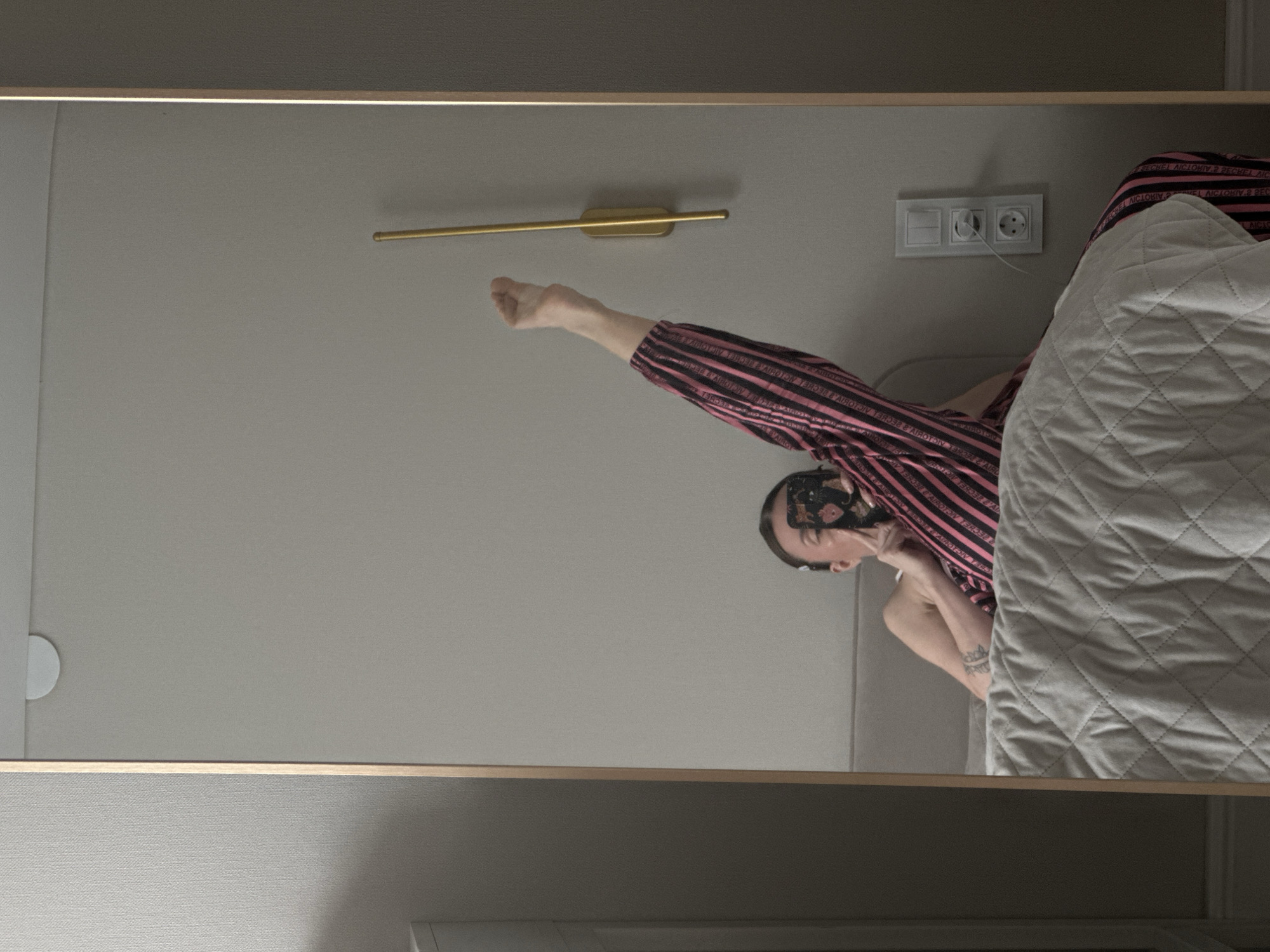 A person is reflected in a large rectangular mirror with a thin gold frame. The person is wearing pink and black striped pajamas and holding a phone with a floral case in front of their face. Their left arm is bent, and their right arm and leg are extended upward toward a gold wall sconce. A white, quilted blanket is visible next to the person. On the wall behind the person and the sconce is a white electrical outlet. The mirror is reflecting an interior space with light gray walls. Based on the reflection of the room, it appears the image was taken in a bedroom or hotel room in Votkinsk, Russia.