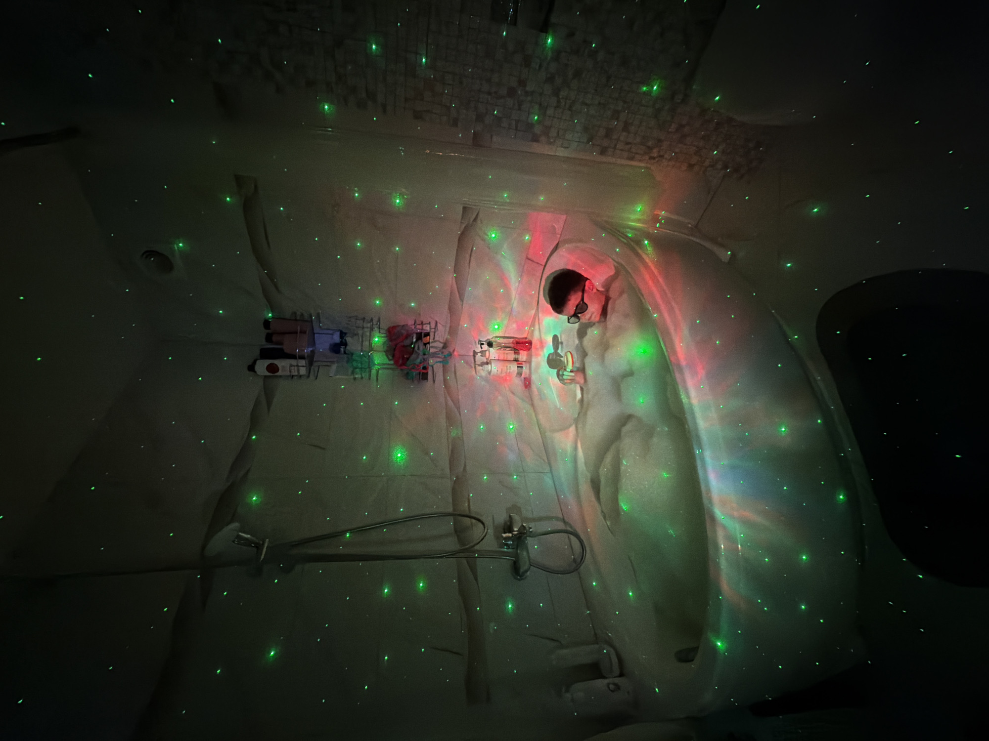 A low-angle, slightly distorted photograph depicts the interior of a dimly lit shower with green and red laser lights scattered across the surfaces. A person wearing eyeglasses and dark hair is partially visible in the background, near the upper right of the image. Various toiletry bottles and dispensers are attached to the wall on the left. A shower head and faucet are centrally located. The walls appear to be constructed of a light-colored material. The scene appears to be a residential bathroom, and the location in the city of Votkinsk, Russia cannot be confirmed from the image.