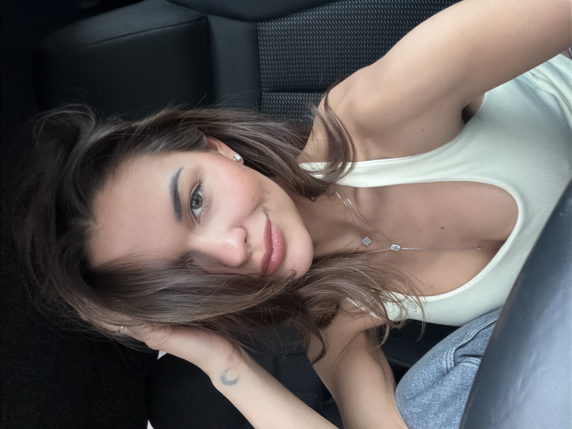 A young woman is shown in a selfie, taken inside a car, possibly in Krasnoyarsk, Russia. She is reclining in the passenger seat, her body angled towards the camera. She has brown hair, fair skin, and is wearing a light-colored tank top, jeans and a necklace with a clover charm. A small crescent moon tattoo is visible on her inner arm. The car has black interior.