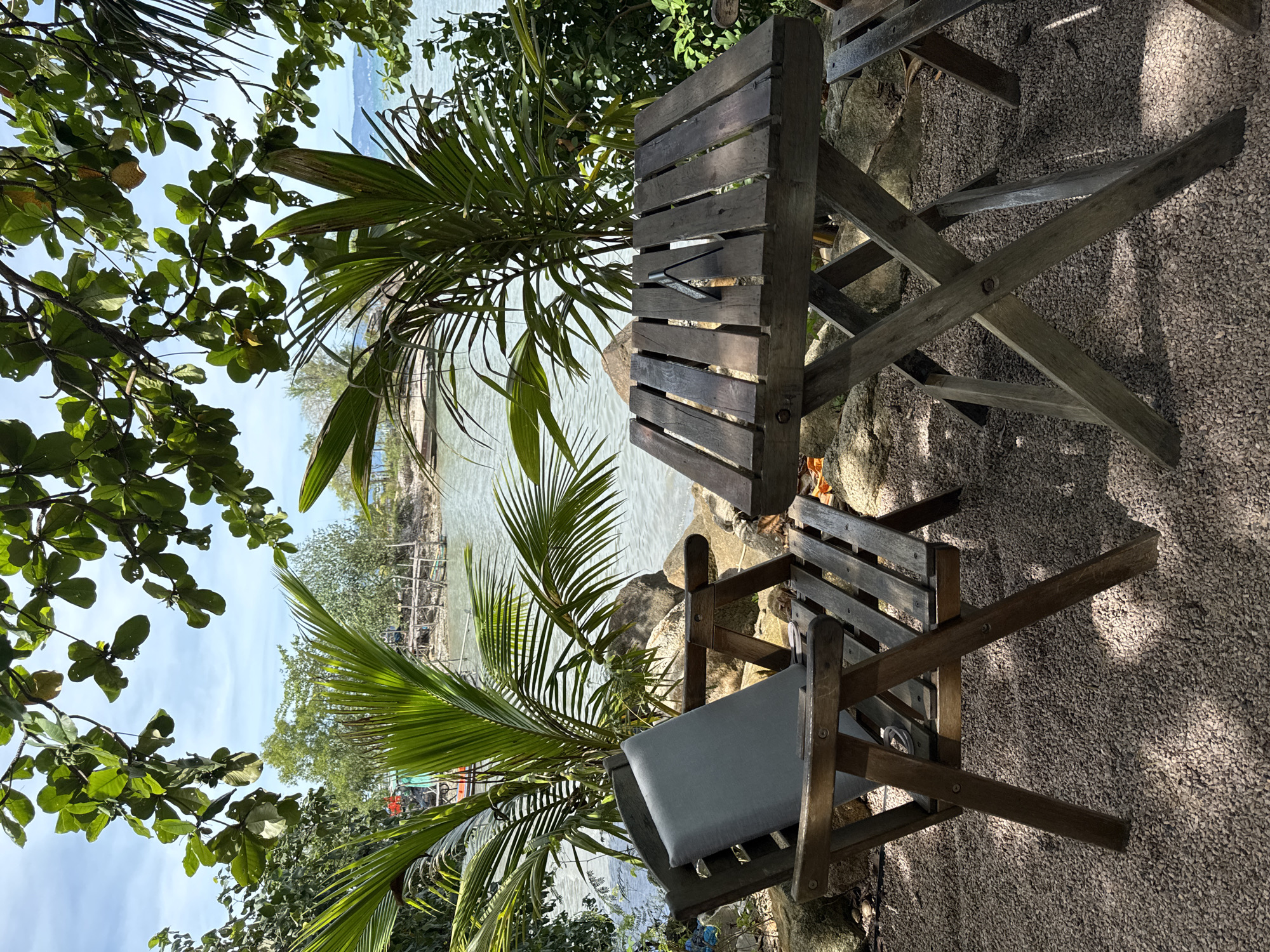 The image depicts a rustic wooden table and chair set in an outdoor setting. The chair has a gray cushion. The area is partially shaded by trees, including palm trees. In the background, there is a view of the sea or a large body of water with a pier or dock structure visible. The ground is covered in small stones or gravel. The location is in Baan Tai, Thailand.