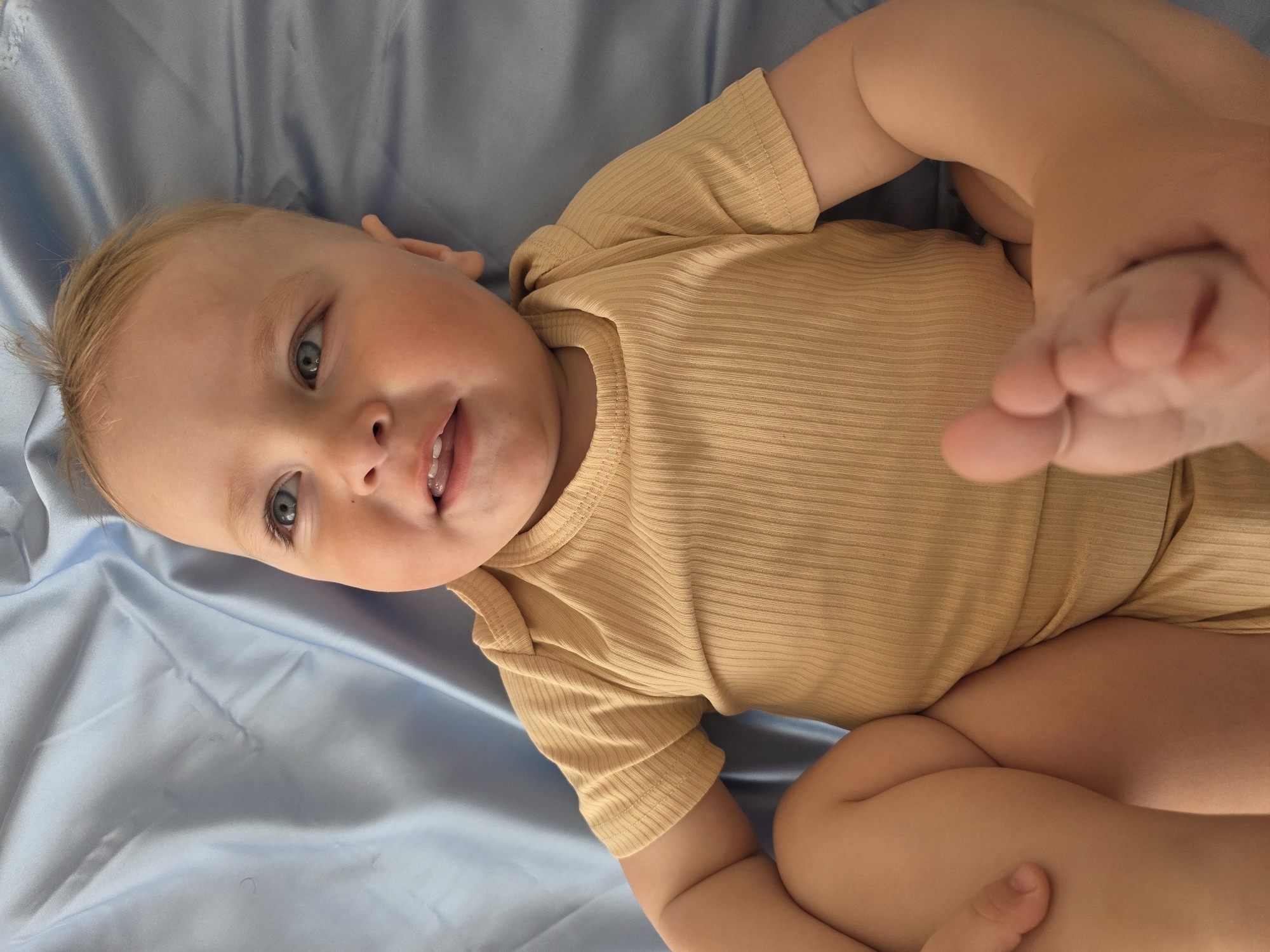 A baby, wearing a beige ribbed one-piece outfit, lies on their back on a blue sheet. The baby is holding their right foot with their hands. The location is not discernible from the image.