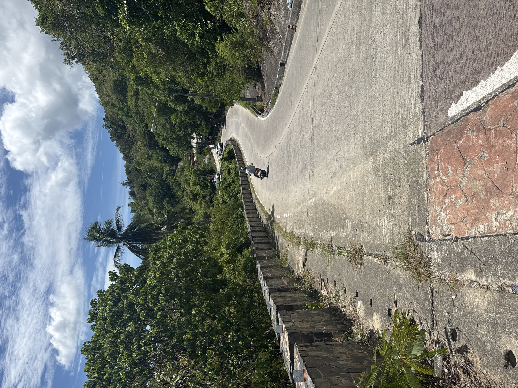 The image depicts a winding asphalt road on a tropical hillside, likely in the Ban Tai Subdistrict of Thailand, characterized by lush green vegetation and a bright blue sky with scattered clouds. The road curves to the right, leading uphill. On the left side of the road, a low stone retaining wall borders the uphill slope, which is covered in dense foliage and trees, including a prominent palm tree.
A lone motorcyclist, wearing a light-colored shirt and dark pants, is riding away from the camera on the road. Further up the road, several cars are visible, indicating ongoing traffic. The road surface appears to be made of concrete in some sections, with visible expansion joints and some areas of wear. The right side of the road slopes downwards, with more dense vegetation. A street lamp is positioned along the side of the road on the right. The foreground shows a section of the road with a cracked concrete surface and a patch of reddish, patterned concrete. Weeds and dry grass are growing along the edges of the road and wall.