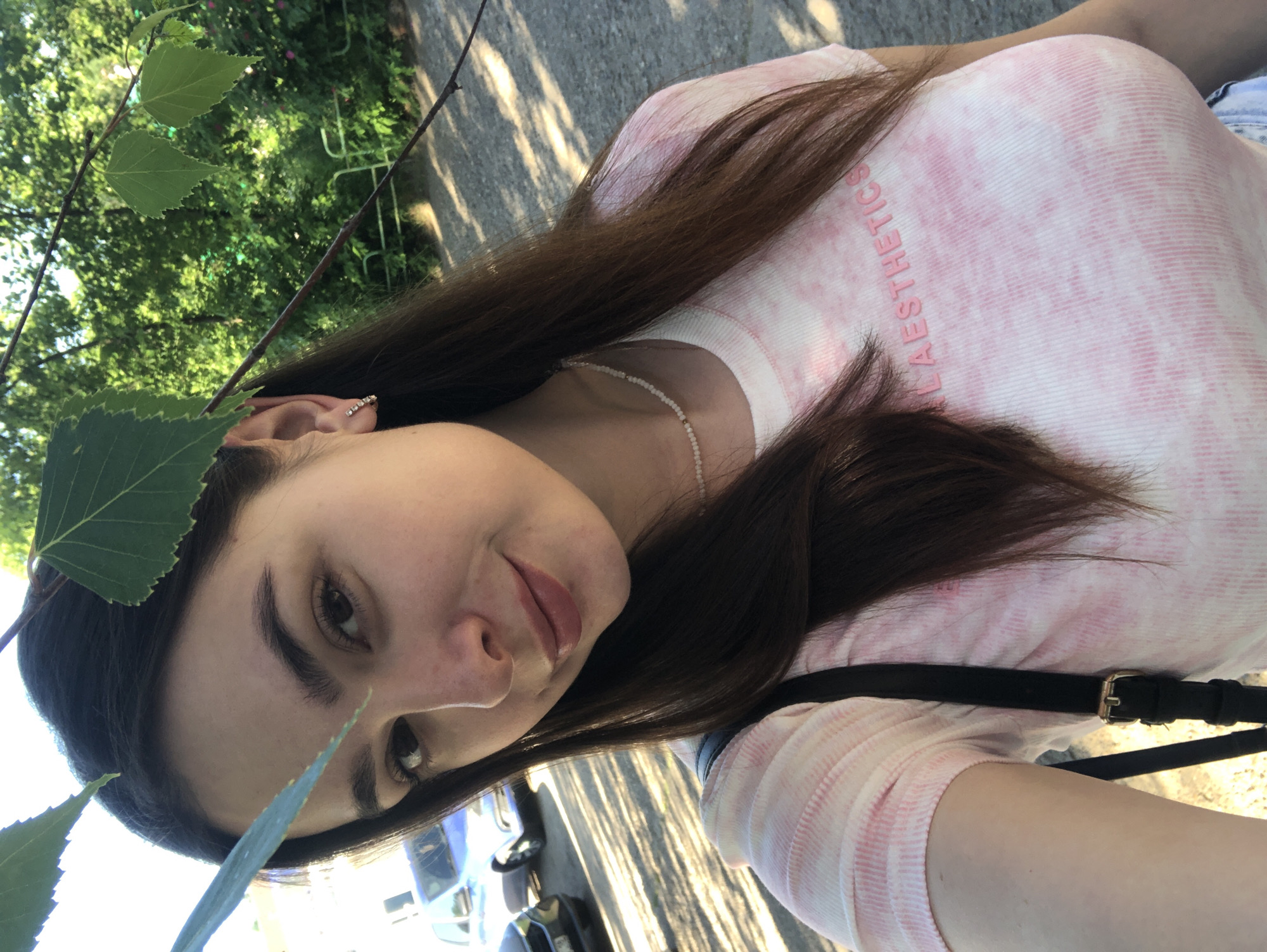 In Noviy, Russia, a young woman with dark hair and fair skin is taking a selfie outdoors. She is wearing a pink and white tie-dye t-shirt with the word "AESTHETICS" printed on it in pink lettering. She also has a delicate pearl necklace. The background shows a tree with green leaves in the foreground, and a parking lot with parked cars and trees in the distance. The sun is shining, casting shadows on the ground.