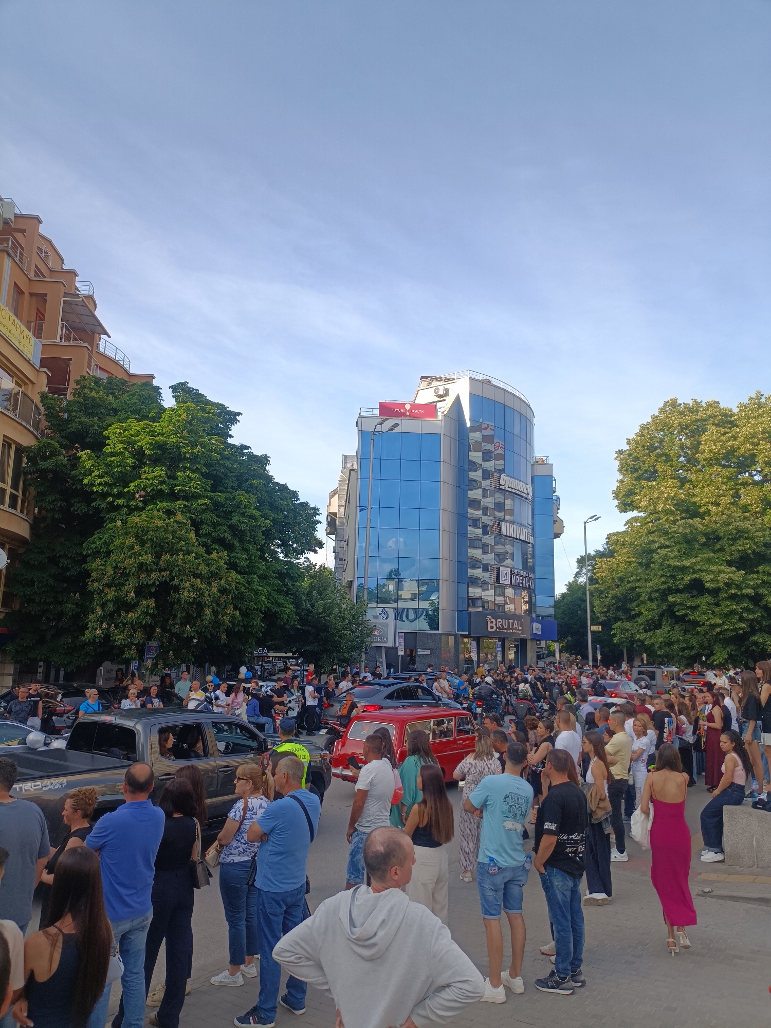 The photograph captures a street scene in Plovdiv, Bulgaria, filled with a crowd of people gathered along the road. A mix of pedestrians and vehicles are present, suggesting a busy urban environment. People are standing, talking, and observing, with some sitting on a low wall. A variety of cars and motorcycles are visible within the crowd. Traffic appears to be congested. The scene is framed by buildings and mature trees, contributing to a mix of urban and natural elements. Street signs and advertisements are affixed to the buildings. The "BRUTAL" brand store is visible as is a building sign for "FUTURE HEALTH". The sky is blue with light clouds.