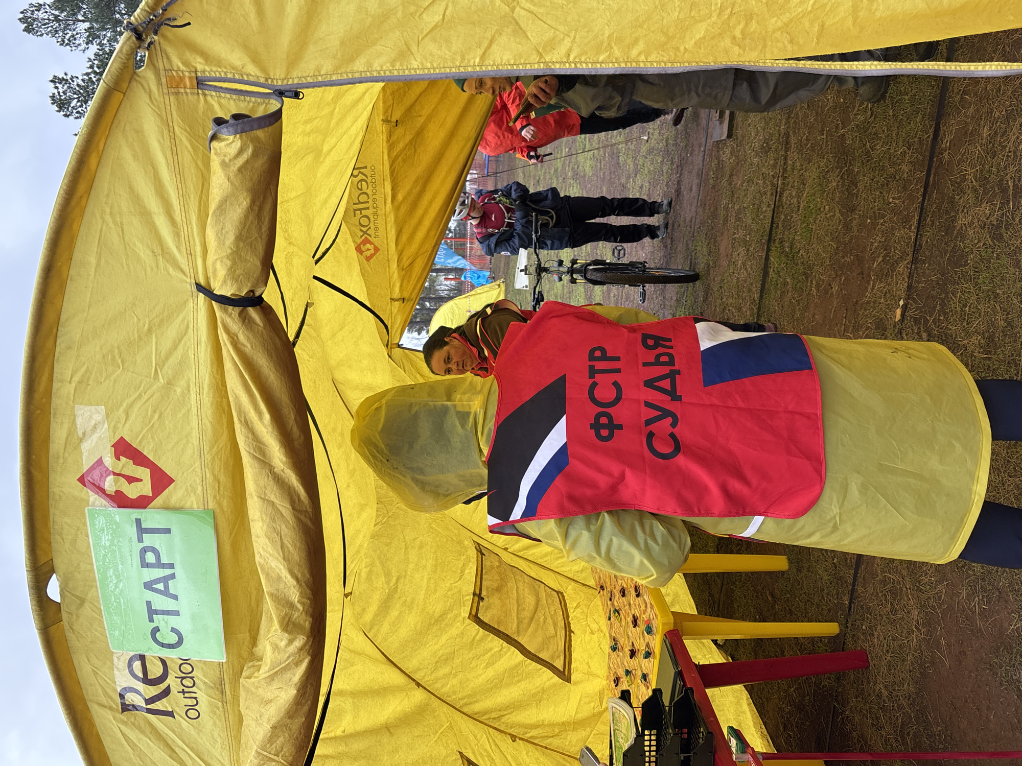 A yellow tent is set up in an outdoor setting, possibly in the city of Orenburg, Russia, based on the lettering on a red vest worn by an individual. The tent is marked with the word "СТАРТ" (START) and an associated logo. A person wearing a red vest with Cyrillic lettering stands near the tent. Another person is visible within the tent, partially obscured. Outside the tent, other individuals are present, with one person on a bicycle. The ground appears to be grassy, and the sky suggests overcast weather.