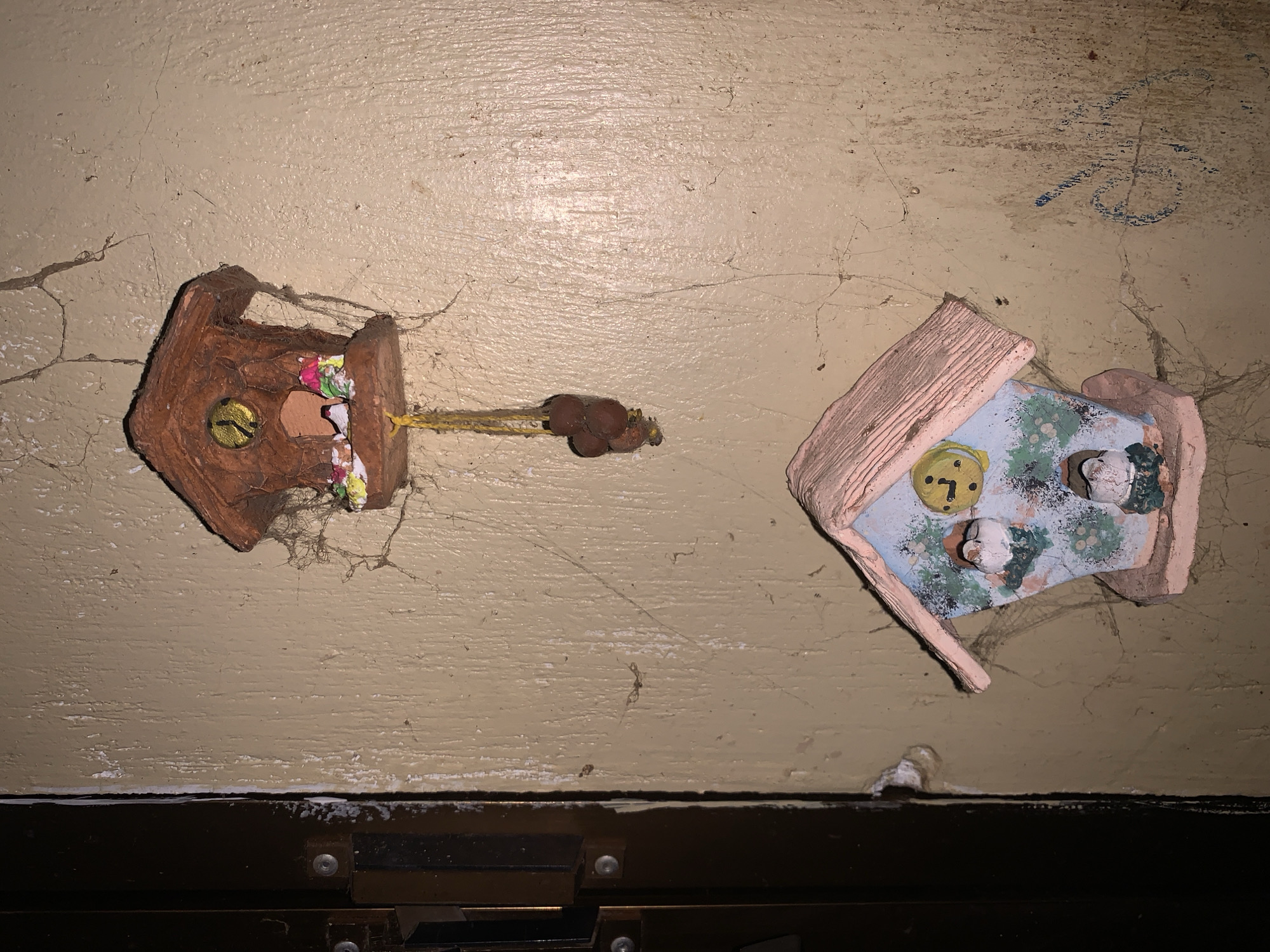 The image presents an overhead or top-down view of two small, decorative house-like objects, positioned against a textured, light-colored background that appears to be an aged wall or ceiling. The surface is marked by fine cracks, dust, and numerous cobwebs, suggesting an old or neglected indoor setting.
On the left, there is a miniature, rustic-looking house crafted from brown material, possibly clay or terracotta. It features a circular, gold-colored embellishment on its front, resembling a button or a bell. The interior of this house is filled with a colorful assortment of soft materials, possibly shredded fabric or paper. A yellow string or cord extends from its right side, adorned with several small, dark brown, spherical objects that could be beads or dried seeds.
To the right, a second miniature house is visible. This structure is lighter in color, possibly light pink or pale beige, and has a light blue interior. The interior is decorated with a green patterned motif, suggestive of foliage or small trees, and includes several small, white, sculpted figures that might represent gnomes or mushrooms. A yellow circular object with dark markings, possibly a clock face or a stylized face, is also visible inside.
In the upper right portion of the background, a faint blue marking appears as an illegible scribble or an abstract symbol. The bottom of the image reveals a dark horizontal band with visible silver screws, indicating a structural element such as a door frame, beam, or part of a piece of furniture. The scene suggests a static display of decorative items within a somewhat aged environment in Santa Ana, Venezuela.