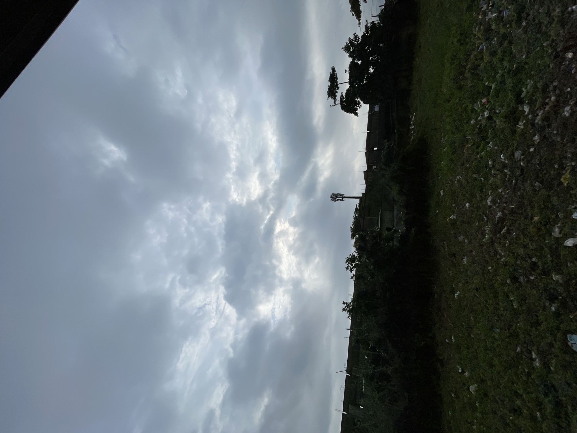 The image captures an overcast sky above an urban-fringe landscape in Ikorodu, Nigeria. The upper two-thirds of the frame is dominated by a dense, gray cloud cover, with diffuse, brighter light breaking through in central and right sections, suggesting a heavily clouded day. Below the horizon, a dark, silhouetted structure, possibly a building or wall, spans the midground, partially obscured by dark, overgrown vegetation. A tall, slender cellular communication tower stands prominently to the left of the frame, also in silhouette. To the right, a cluster of dark-foliaged trees with distinct, somewhat flat canopies is visible. The foreground consists of an expansive, uneven green field covered in short, dense vegetation. This green space is significantly littered with various forms of refuse, including numerous white and light-colored plastic bags, wrappers, and other debris scattered throughout. No people, animals, actions, or interactions are present in the scene. A dark, diagonal obstruction is visible in the upper right corner, indicating the vantage point of the photographer.