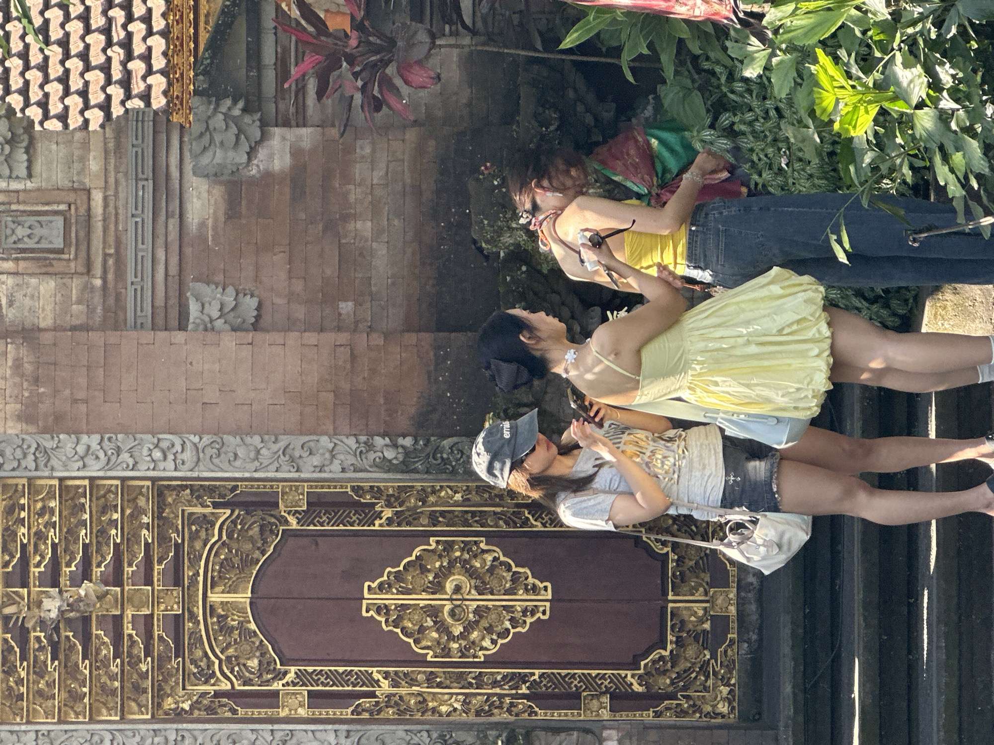 In the city of Ubud, Indonesia, three young women stand near a temple entrance. The woman on the left, wearing a gray t-shirt, denim shorts, and a baseball cap, is looking at her phone. She carries a white purse. In the center, a woman in a yellow dress holds a phone and appears to be taking a picture. To her right, a third woman, wearing a yellow top and blue jeans, is also looking at her phone, and another woman in traditional clothing is partially visible behind her. The backdrop features a detailed wooden door with gold accents, a brick wall, and lush green foliage. The scene is bathed in bright sunlight, casting shadows on the stone steps.
