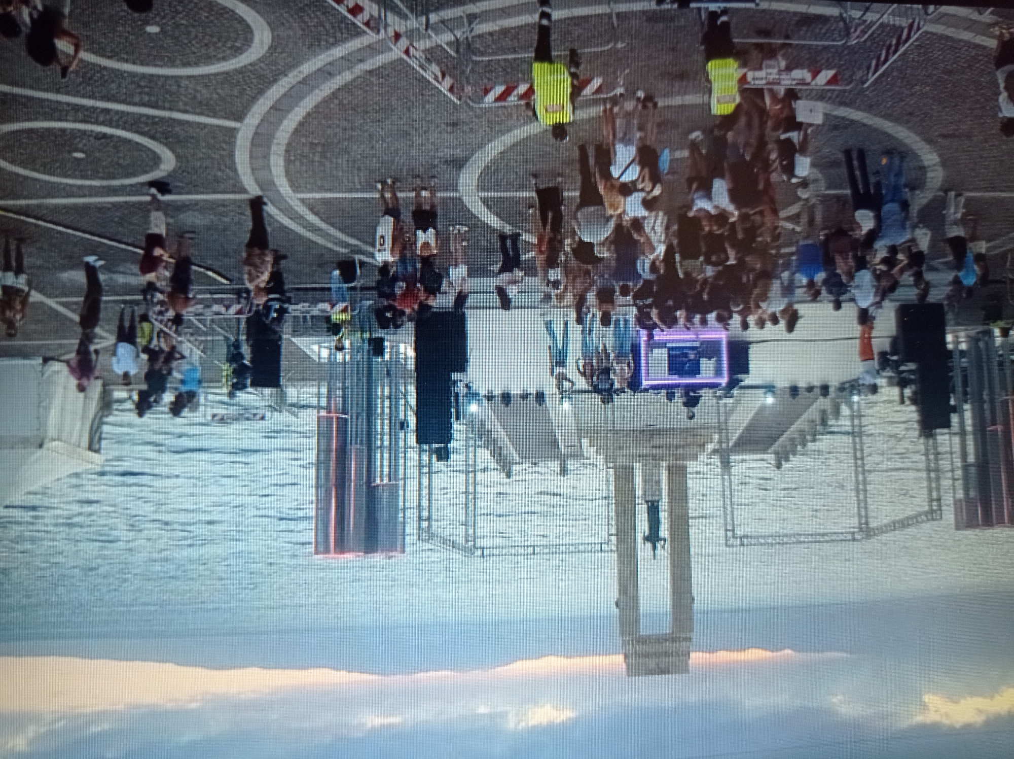 The image depicts an outdoor event with a crowd of people gathered near a body of water, likely a lake or sea. In the background, there is a large monument with two pillars and a statue in the center, overlooking the water. A stage or platform with scaffolding and lighting equipment is set up in front of the monument, suggesting a performance or concert. Several people are visible on the stage, possibly performers or technicians. The foreground shows a plaza with a patterned pavement, where many spectators are standing and mingling. Some individuals are wearing bright clothing, and there are barriers set up in some areas, indicating crowd control. The sky is overcast with some hints of sunset colors. The scene appears to be a lively public gathering in a scenic location. The provided image and crops do not contain any information to suggest the location is the city of Mukachevo, Ukraine.