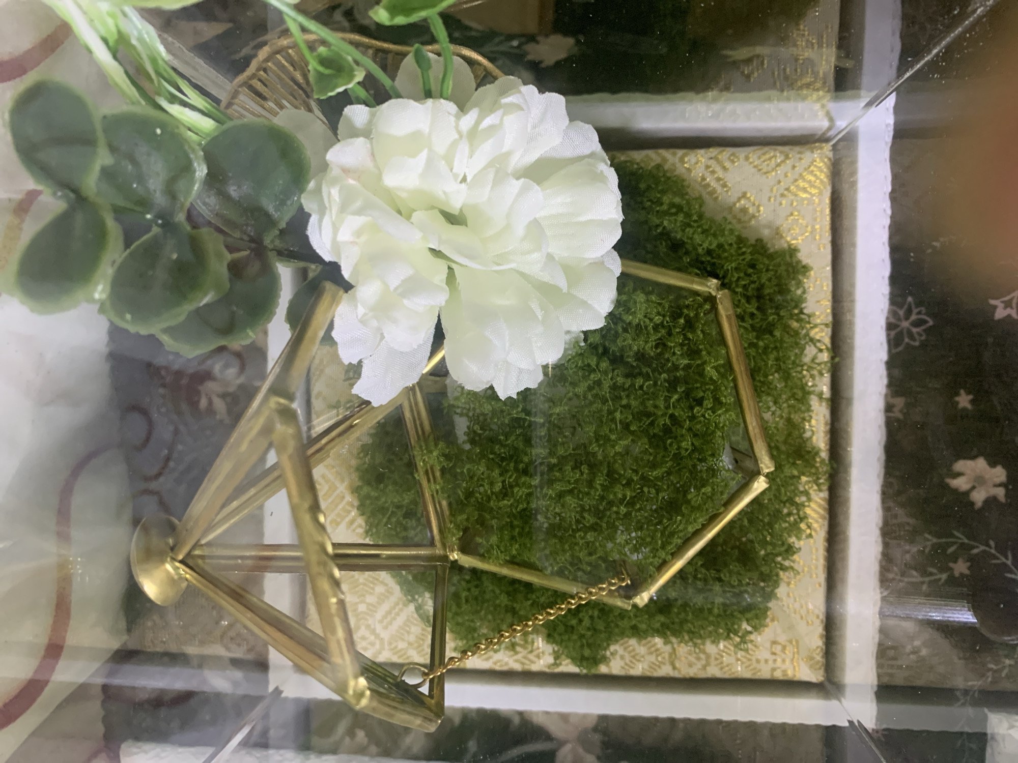 The image shows a decorative item inside a glass container. The item consists of a geometric brass frame with a gold chain, filled with green moss. A cluster of white artificial flowers is placed next to the moss, and some green artificial leaves are visible in the background. The container itself appears to be made of glass and has a reflective surface. The background outside the container shows a patterned fabric. The scene is static and does not depict any people, actions, or interactions. The location in Kuala Pembuang, Indonesia, is not discernible from the visual information in the image.