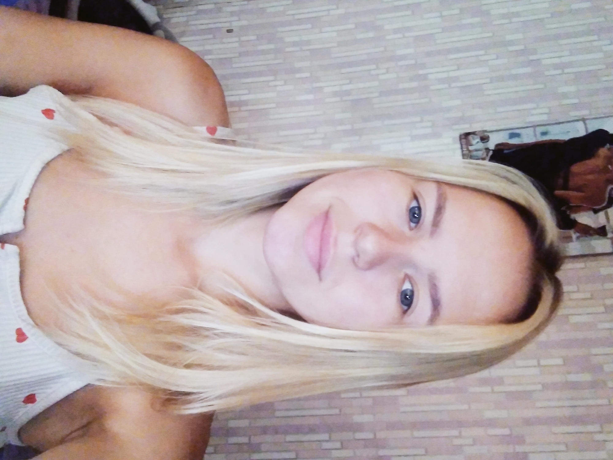 A young woman with blonde hair and blue eyes is looking directly at the camera, her head slightly tilted. She is wearing a white tank top with a subtle pattern. The background appears to be a wall with a textured wallpaper. On the left side of the frame, a framed picture is partially visible. The image is a close-up, focusing on the woman's face and upper body. No specific location within Radisne, Ukraine, can be identified from the visual information provided.