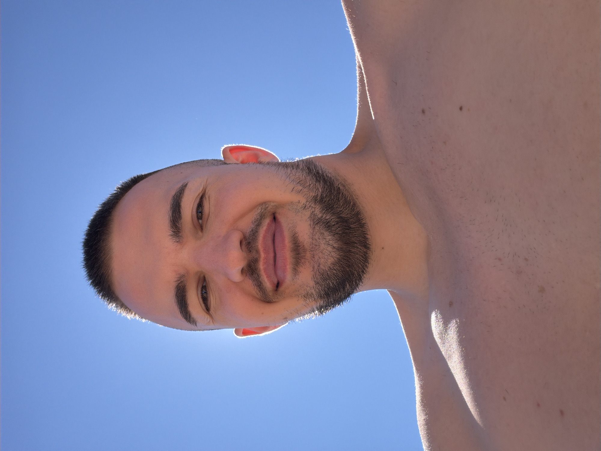 A low-angle, close-up selfie is taken of a fair-skinned man with dark hair and a short beard. He is outdoors against a bright blue sky, with the sun positioned behind his head, creating a halo effect around his hairline and ear. The man is looking directly at the camera with a slight smile, his eyes are open, and his lips are closed. His shoulders and upper chest are visible and appear bare. The man's skin is lightly tanned and shows some minor blemishes. The sky is clear and cloudless. The image was captured in Primorsko, Bulgaria.