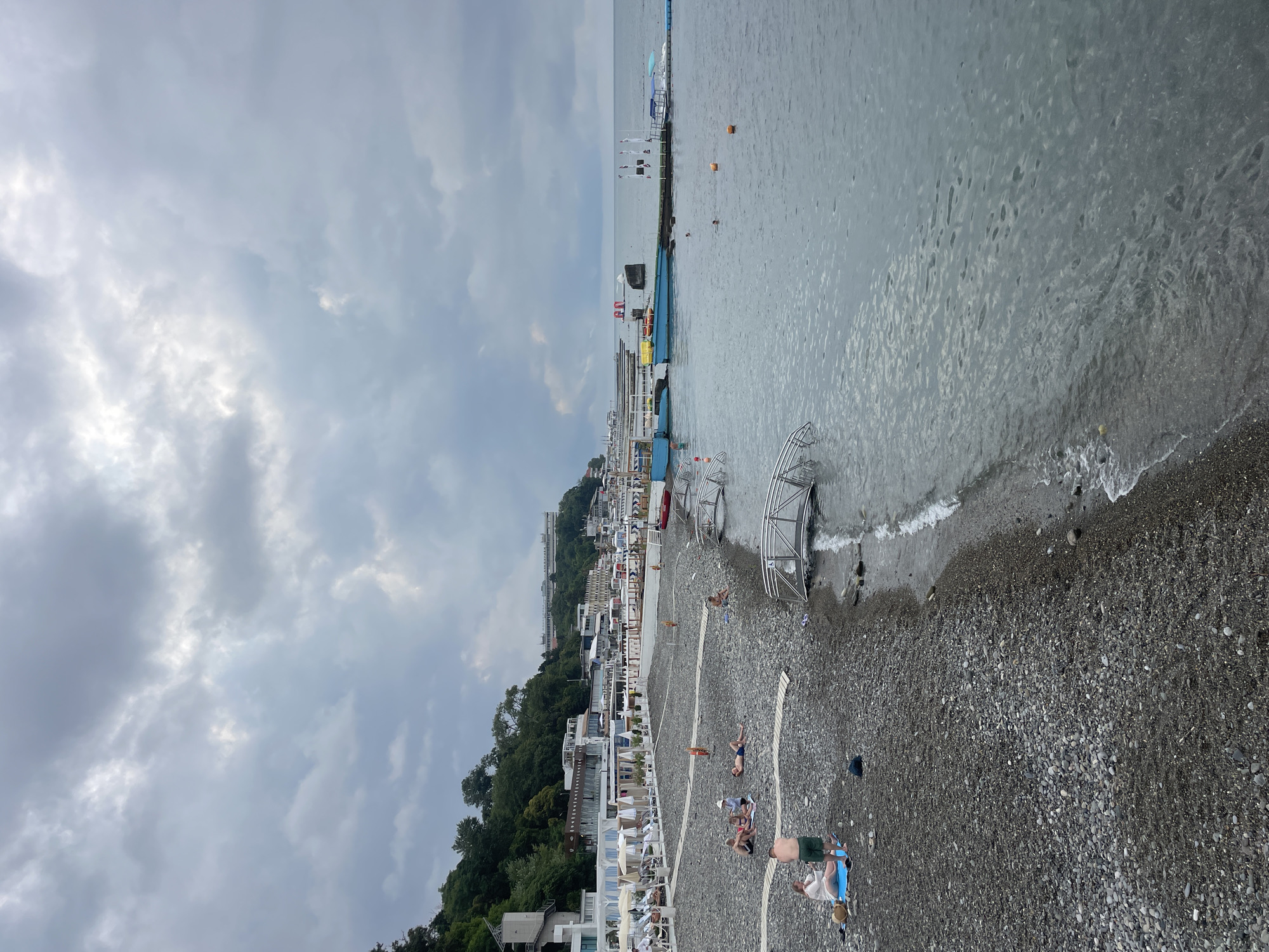 The image depicts a cloudy day at a pebbled beach in Sochi, Russia. The beach is lined with white umbrellas and lounge chairs, suggesting a resort area. Several people are visible on the beach, some sunbathing and others sitting. In the water, there are several boats and a pier with a few individuals swimming. The sea is calm with gentle waves lapping the shore. The background shows a hillside covered in dense green trees, with buildings nestled amongst them, hinting at the urban sprawl of Sochi extending up the slopes. The overall scene conveys a tranquil, albeit overcast, seaside atmosphere.