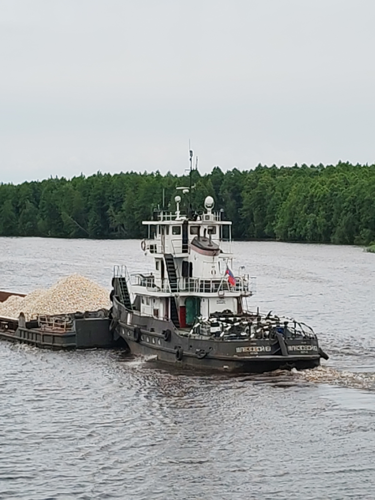 This media file shows a tugboat pushing a barge laden with a light-colored, granular material, likely stone or gravel, down a river. The tugboat is dark gray with a white superstructure and a prominent cabin. A Russian flag is visible on the tugboat's superstructure. The barge is long and rectangular, filled high with the cargo.
The scene is set on a wide river with calm water, creating gentle ripples. In the background, a dense, green forest lines the riverbank, suggesting a natural, possibly remote, location. The sky is overcast, indicating a cloudy day, and there are no strong shadows. The lighting is diffused.
The overall impression is one of industrial activity within a natural landscape. The tugboat and barge are the primary objects, engaged in the transportation of goods. The lack of visible people adds to the focus on the vessels and the task. The setting appears to be a waterway in Novokemskii, Russia, given the location context.
Visible text on the stern of the tugboat appears to be in Cyrillic script. It reads "ВЛЕСЕВИЧ" on the left side and "ВЛЕСЕВИЧ" again on the right side, likely the name of the vessel.