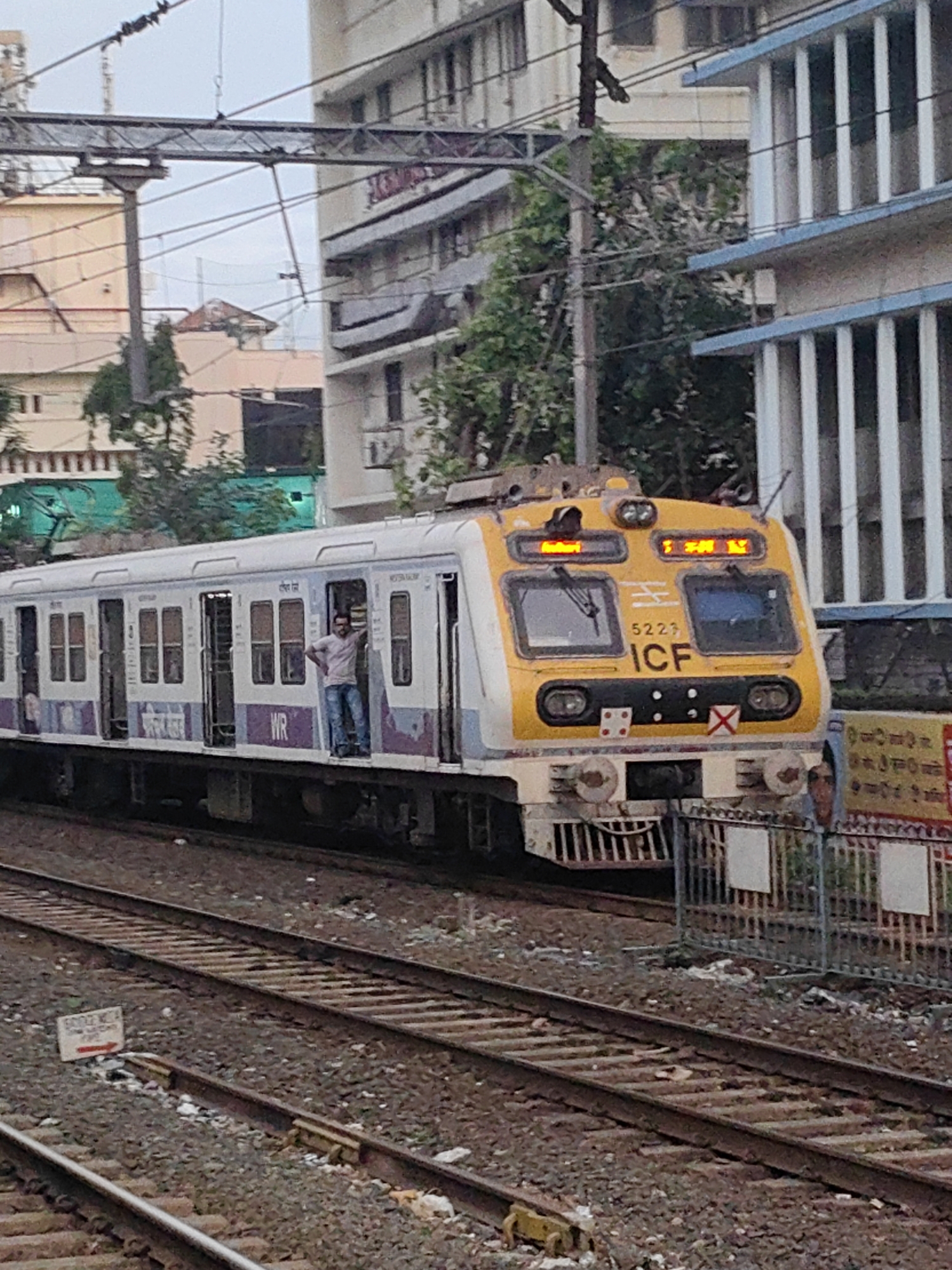 A train with a yellow and white front is stopped at a station in Mumbai, India. A man is standing in the open doorway of the train. The train cars are white with purple and blue accents, and display the letters "WR" and some text in an Indian script. The train station platform is visible, with overhead wires and power lines. In the background, there are buildings, some of which appear to be residential or office buildings, and trees. The tracks in the foreground are made of gravel and ballast, and there are several sets of parallel tracks. A white sign with an arrow pointing to the right is visible on the ballast near the tracks. The overall scene depicts a typical urban train station environment in Mumbai.