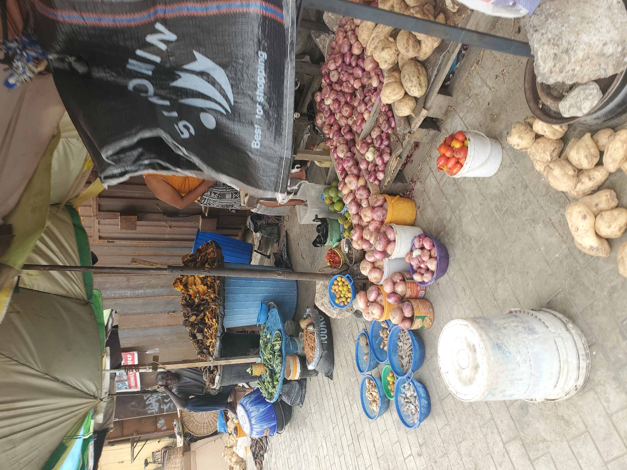 This image captures a bustling outdoor market stall, likely in Lagos, Nigeria, teeming with fresh produce and various goods under natural daylight. The ground is paved with light-colored interlocking bricks.
Prominently displayed are numerous red onions, piled high on wooden crates and overflowing from various plastic buckets (yellow, white, and purple). Adjacent to these, large quantities of yams or sweet potatoes are stacked on wooden pallets and directly on the ground. A white bucket holds ripe red tomatoes, while other containers feature green mangoes or avocados, and yellow citrus fruits such as lemons or limes.
Further within the stall, several blue plastic baskets are filled with small dried fish or seafood. Green leafy vegetables and okra are also visible on display, some within blue buckets. Sacks labeled "100KG" suggest the sale of grains or beans. A weighing scale with a large stone weight is visible near some yams.
A large black tarpaulin, serving as a canopy, features the white text "NINGIS" with a logo and the slogan "Best for shopping." Partially visible behind this tarpaulin is a person wearing an orange top. On the left side of the stall, a dark-skinned man in a grey shirt and dark trousers sits under a beige and green umbrella, appearing to be a vendor. A red sign with "747" is visible behind him. The market infrastructure includes wooden planks and metal poles, supporting the various displays and shelters. The scene portrays a typical vibrant market environment with diverse food offerings.