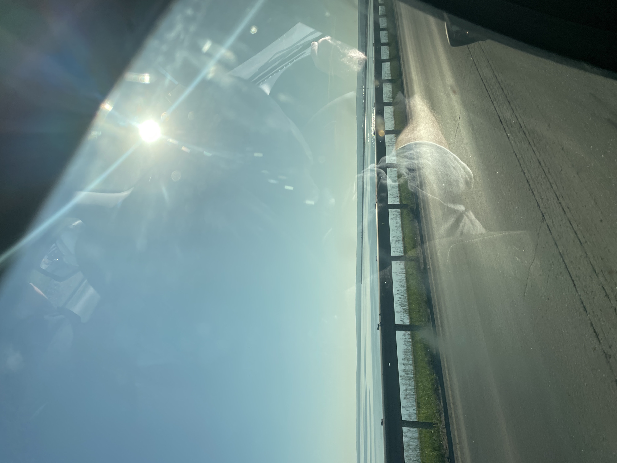 The image is a snapshot taken from inside a vehicle, looking out through the windshield. The sun is bright and positioned in the upper right quadrant, causing lens flare and reflections on the glass. The scene outside depicts a body of water, likely the Kama River or its reservoir, stretching to the horizon under a clear blue sky. A guardrail runs parallel to the road in the mid-ground, separating the asphalt from a grassy embankment. The road surface is visible in the foreground, showing tire marks and some debris. The reflections on the windshield are prominent, obscuring some of the details of the external environment, and include what appears to be the interior of the car and potentially a person's hand. This view is consistent with a road near the waterfront in Chaikovsky, Russia, possibly along the highway leading to or from the city, given the expansive water body and road infrastructure.