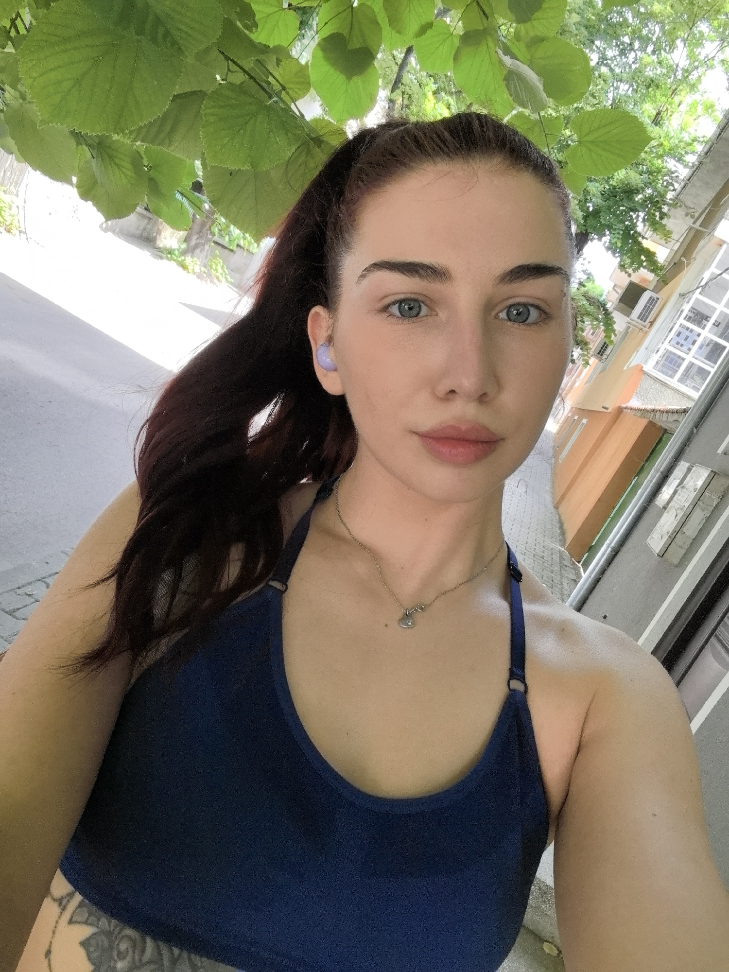 A young woman takes a selfie outdoors on a street in Stara Zagora, Bulgaria. She wears a blue athletic top, and a necklace with a small pendant. Her dark hair is pulled back in a ponytail, and she has a light purple earpiece in her left ear. The background includes a narrow street with sidewalks, buildings with light-colored facades, and leafy trees overhead. The sky is bright.