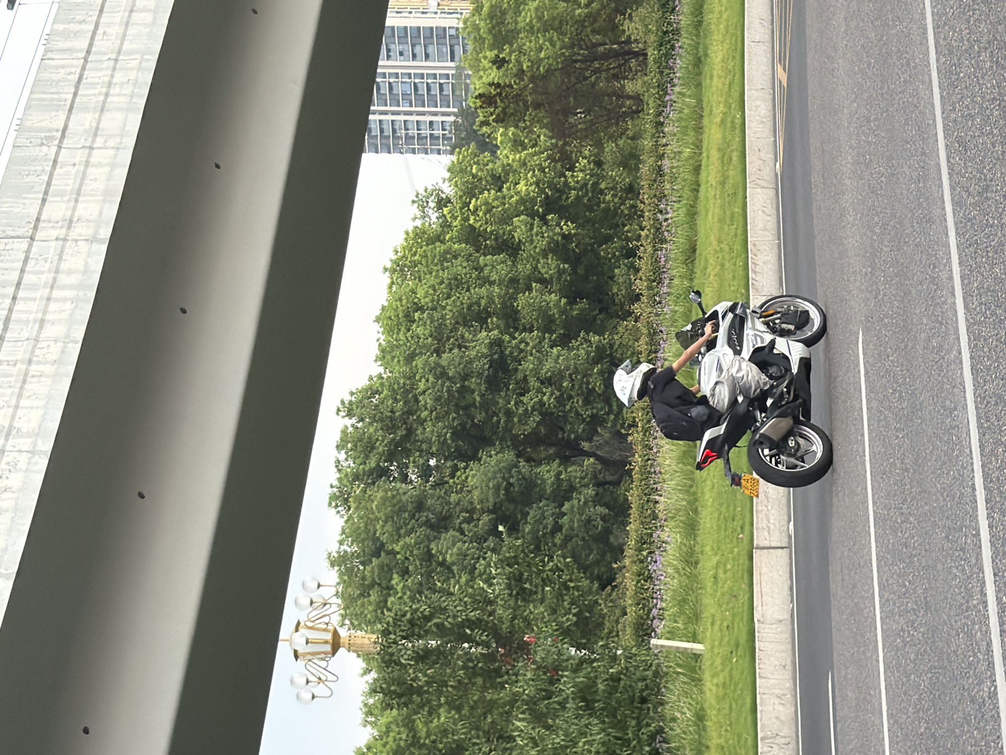 A person wearing a white helmet and a black shirt rides a white motorcycle with a black backpack on a paved road in Jiangjin, China. The motorcycle has a yellow license plate. The rider is in motion, leaning into a turn. The road is lined with a grassy median and a line of trees. In the background, there are tall buildings and a bridge overhead. The sky is overcast.