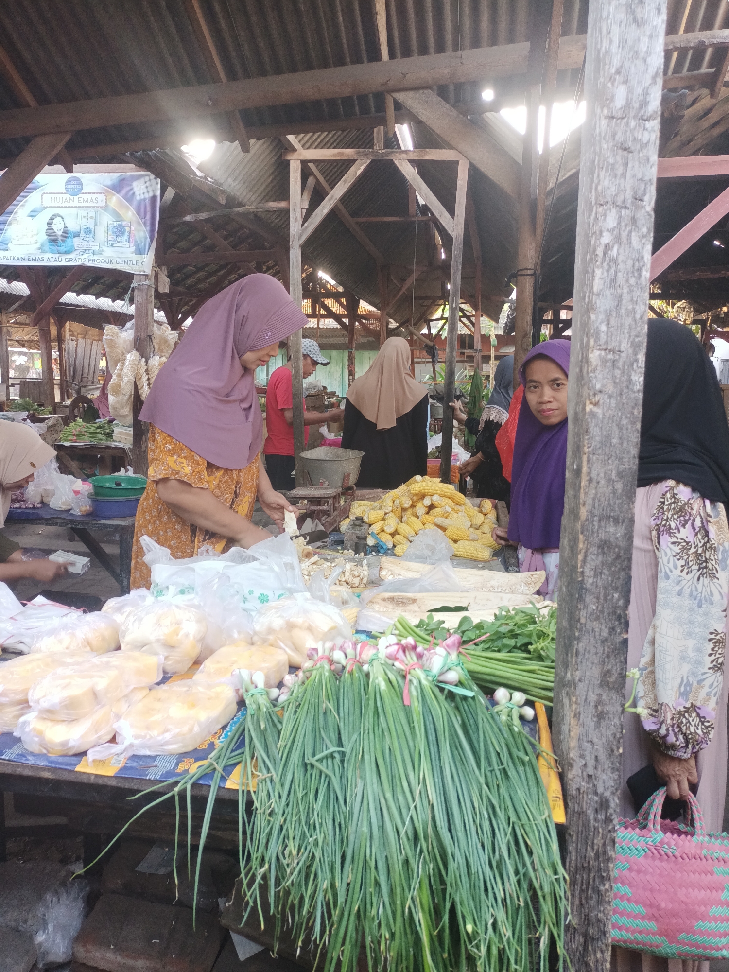 In the city of Badas, Indonesia, a market scene unfolds under a weathered, corrugated metal roof supported by rough wooden beams. Sunlight streams through gaps in the structure, illuminating the various goods and people present.
In the foreground, a stall displays an abundance of fresh produce. Bunches of long, green scallions with pinkish bulbs are prominently arranged, tied together with green string. Behind them, a large pile of yellow corn cobs is visible, some still partially husked. To the left, a selection of white, pillow-shaped items, likely some form of food product, are neatly packaged in clear plastic bags.
Several individuals are engaged in activities at the market. A woman wearing a mauve hijab and an orange patterned dress is the central figure, actively handling food items and appearing to prepare them for sale, possibly wrapping something in plastic. To her left, another woman, also wearing a hijab, is partially visible, seemingly tending to goods on her stall.
Further back, a woman in a beige hijab stands near a stall. To the right of the central figure, another woman, wearing a purple hijab, looks directly at the camera. Behind her, a person in a black hijab and a patterned garment is visible. A man in a red shirt stands in the middle ground, appearing to be a vendor or customer.
The environment suggests a bustling, open-air market. Various wooden structures, stalls, and potentially other vendors and customers are implied by the depth of the scene and the lighting. The overall impression is of a vibrant local marketplace, with the focus on fresh food and the interactions between vendors and shoppers.