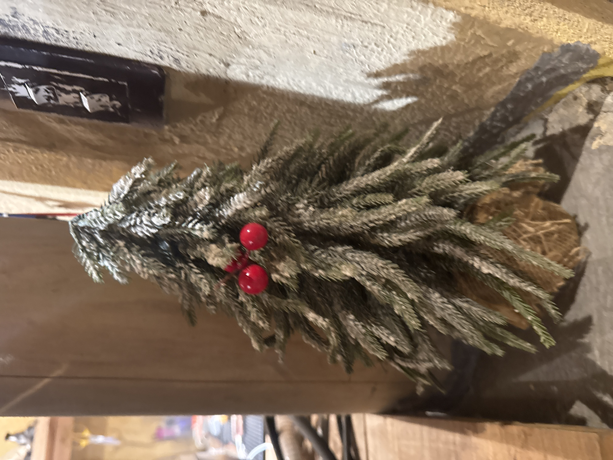 The image shows a small, artificial Christmas tree decorated with red berries, likely placed on a shelf or table. The tree has a snowy or frosted appearance. The background is indistinct but appears to be a wall and other surfaces, possibly within an indoor setting. There is no information to suggest this scene is in Lũng Cú Commune, Vietnam. The image appears to be taken indoors, with the artificial Christmas tree as the focal point.