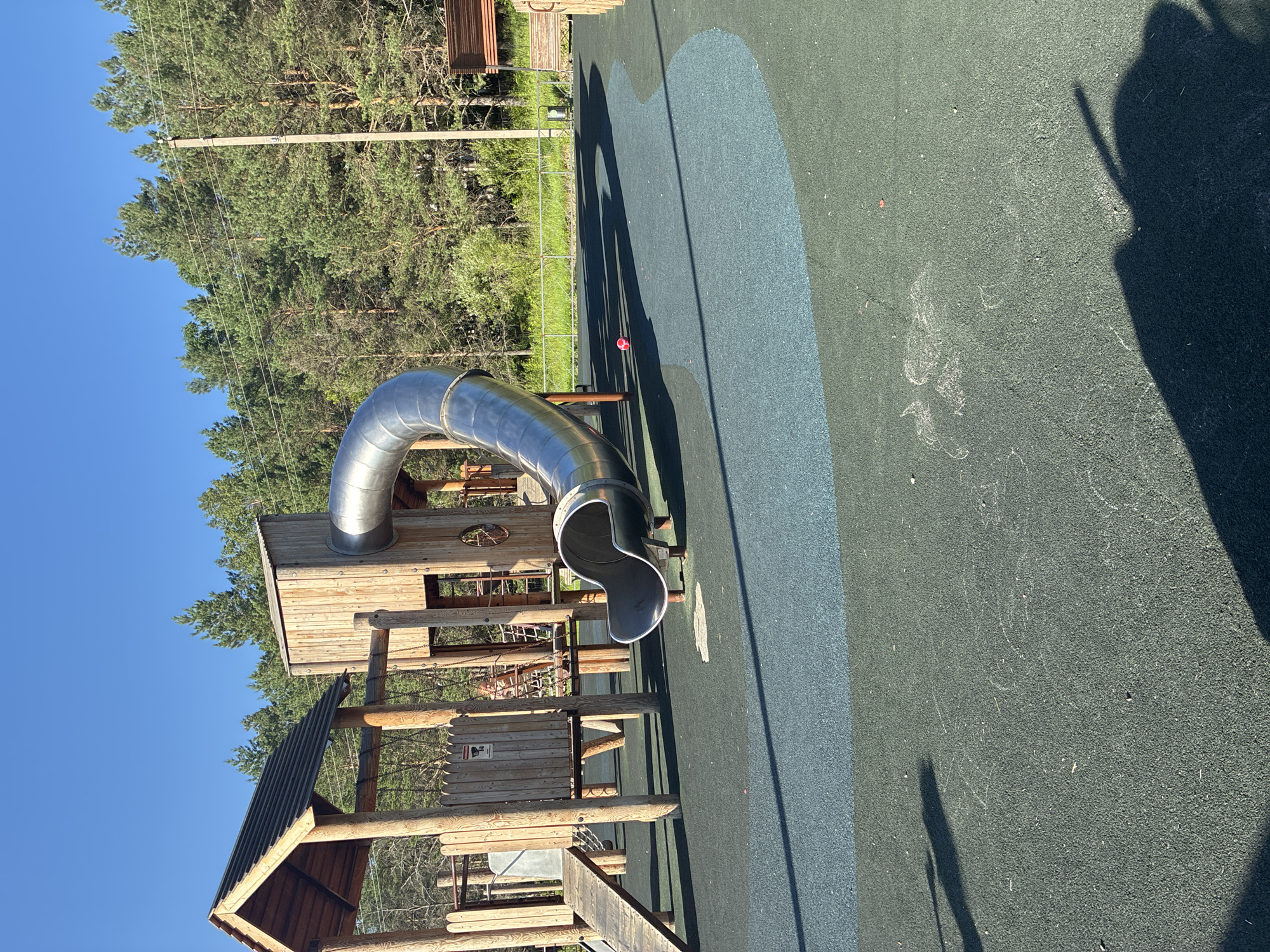 The image depicts an outdoor playground with a prominent metal slide and wooden play structures. The playground surface is a mix of dark green and blue rubberized material. In the background, a dense forest of pine trees lines the horizon under a clear blue sky. A wooden utility pole with wires is visible on the right side of the frame, near the edge of the forest. A small red ball lies on the blue section of the playground surface. The scene is set in the city of Gornye Kljuchi, Russia.