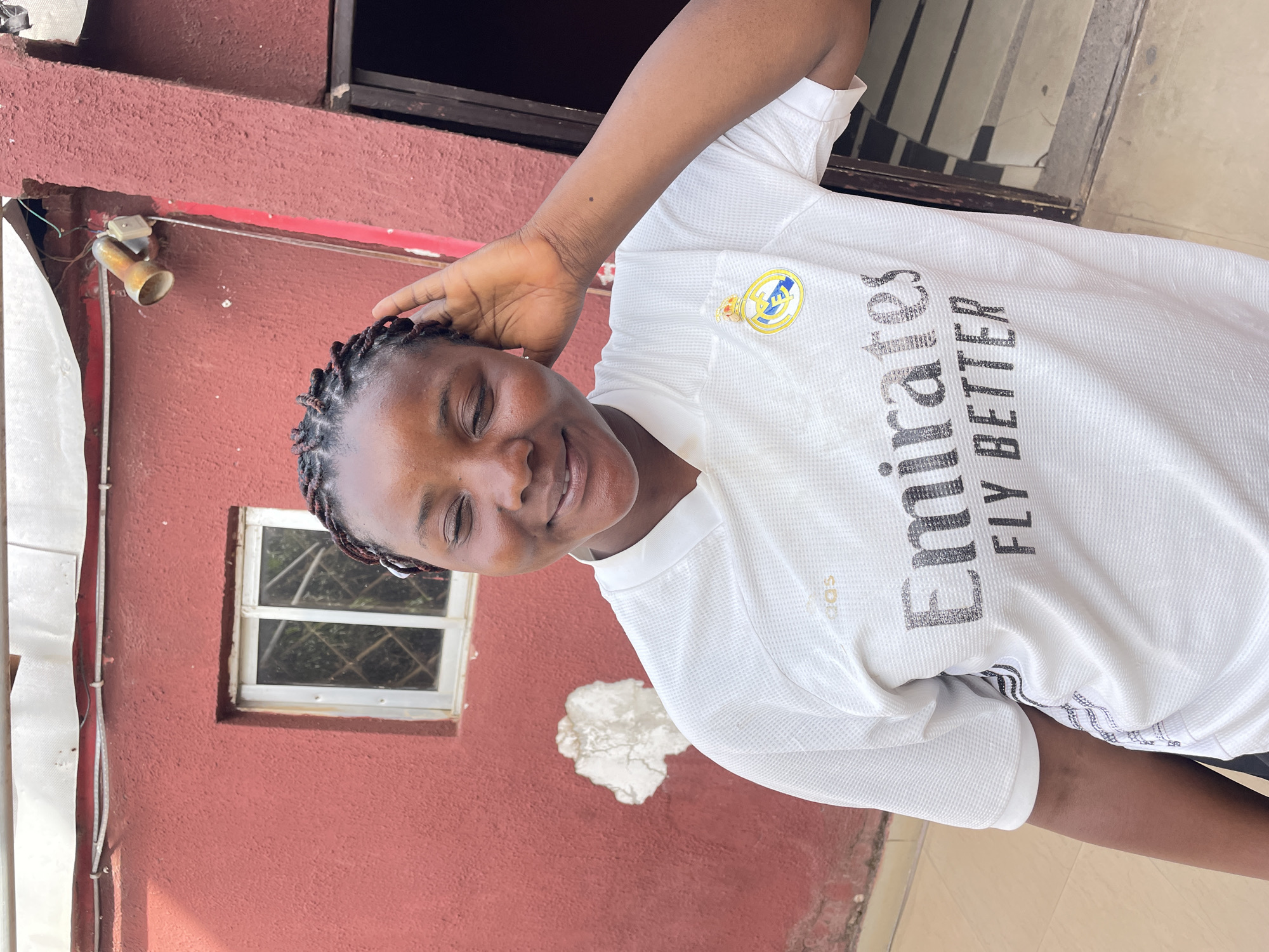 A smiling young woman with dark skin and braided hair poses for a photo, looking directly at the camera. She is wearing a white short-sleeved sports jersey with a textured pattern. Her left hand is raised, with her fingers resting gently near the back of her head.
The jersey prominently features the Real Madrid crest on the left chest and the text "Emirates FLY BETTER" in dark lettering across the front. A smaller "adidas" logo is visible on the right chest area.
The background consists of a reddish-brown, textured exterior wall, suggesting an outdoor or semi-outdoor setting. To the left of the woman's head, an old, disconnected light fixture with visible wiring is attached to the wall. Further down on the left, a white-framed window with a diamond-patterned security grate is visible. A white vertical conduit or pipe runs along the wall to the left of the window. A patch of irregularly shaped white plaster is visible on the wall behind her right shoulder. The lighting is bright, indicating it is daytime. The woman appears happy and relaxed. The architectural elements and overall scene are consistent with a location such as Jos, Nigeria.
