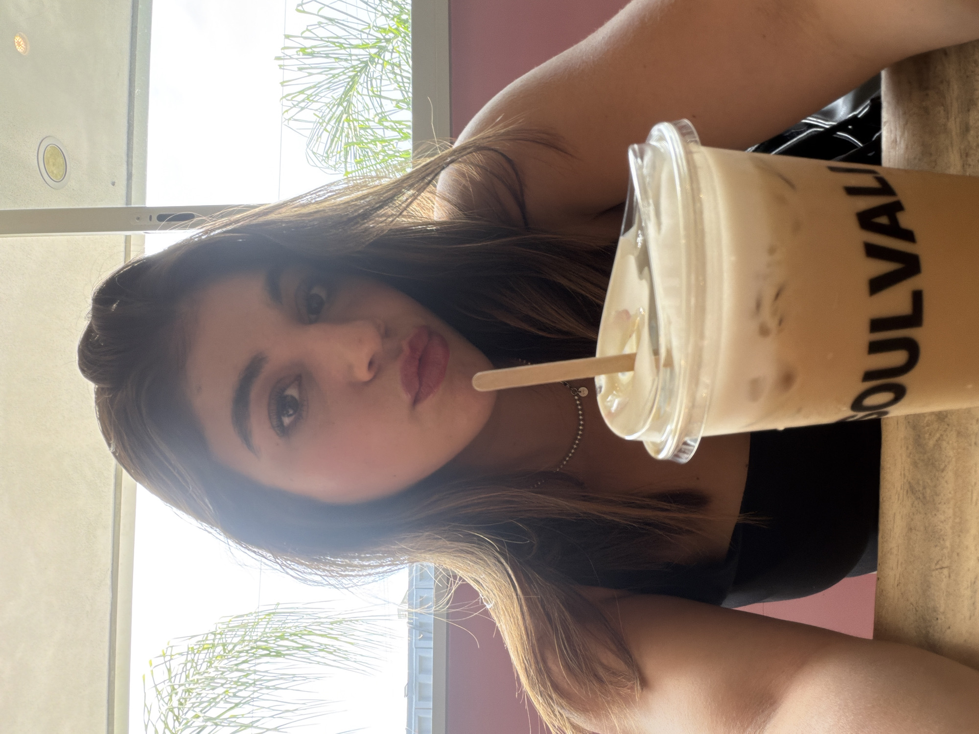 A young woman is pictured in a selfie style shot. She has brown hair and light skin, and is wearing a black top. She is holding a plastic cup of iced coffee with a straw. The cup has "SOULVALL" printed on it in black letters. The background shows a window with palm trees visible outside. The interior appears to be a cafe or restaurant. Given the cup's inscription, it can be inferred that this photo was taken at Soulvalle, a chain of cafes and bakeries in Puebla, Mexico.