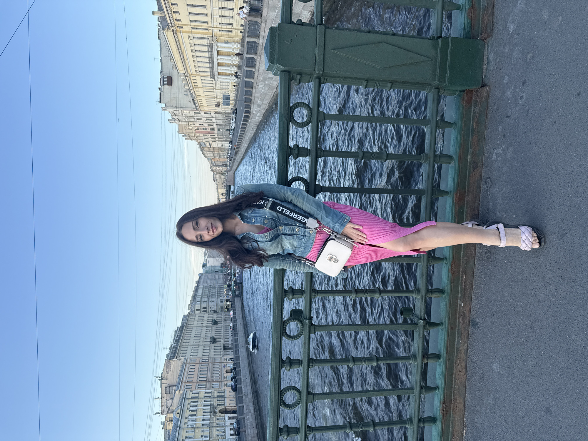 A young woman stands on a bridge overlooking a canal in Saint Petersburg, Russia. She is wearing a denim jacket over a pink dress, and a white crossbody bag. The canal is lined with ornate buildings, and a boat is visible in the distance. The sky is clear and blue. The bridge railing is made of dark green metal with decorative circular elements. The water in the canal is dark and choppy, with visible ripples. The buildings on either side of the canal have a classical architectural style with many windows. There are a few people visible on the far side of the canal walking along the embankment.