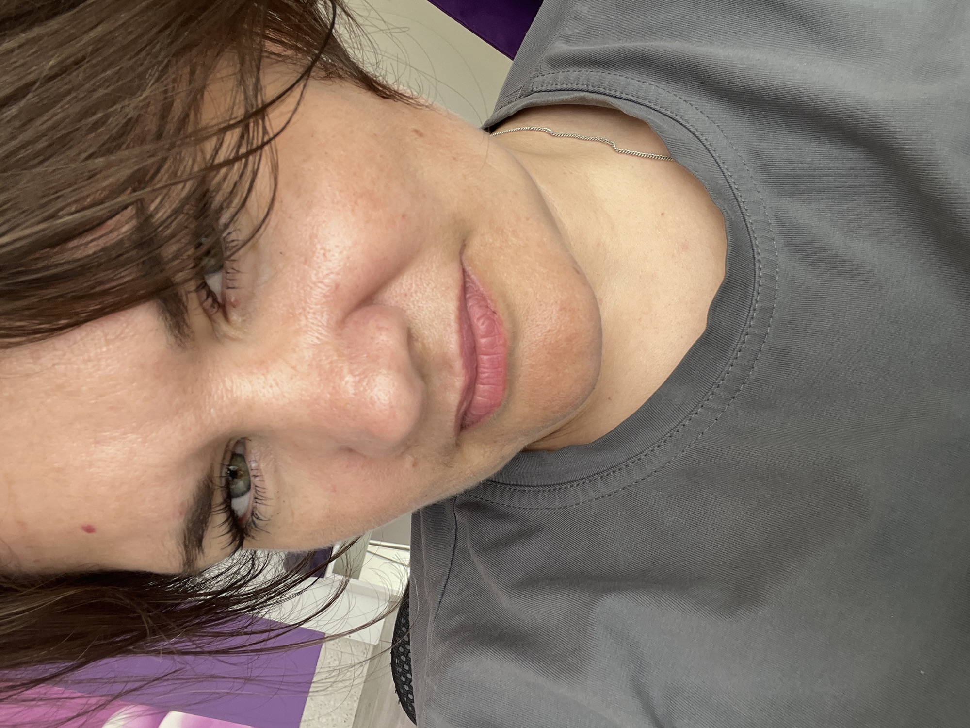 The image is a close-up, self-portrait of a woman with brown hair and green eyes. She is wearing a grey t-shirt and a thin silver chain necklace. Her skin appears fair with some redness and visible pores. There is a small reddish mole on her forehead. The background is blurred, showing hints of purple and white. The image appears to be taken indoors, possibly in Afipsky, Russia, based on the prompt.