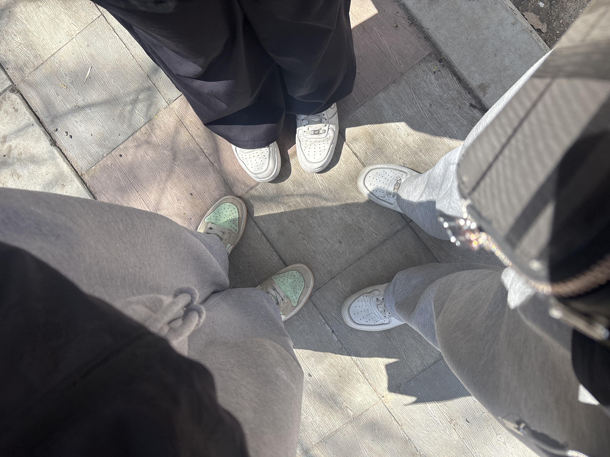 Three people stand on a paved surface, possibly a sidewalk, in Yerevan, Armenia. All individuals are seen from the waist down. The person on the left is wearing light grey sweatpants and muted green sneakers. The person in the middle is wearing wide black pants and white sneakers. The person on the right is wearing grey sweatpants and white sneakers. Their feet are oriented towards the center, suggesting a group gathering. The pavement consists of rectangular tiles with a faux wood grain pattern. Shadows suggest the image was taken during daylight.