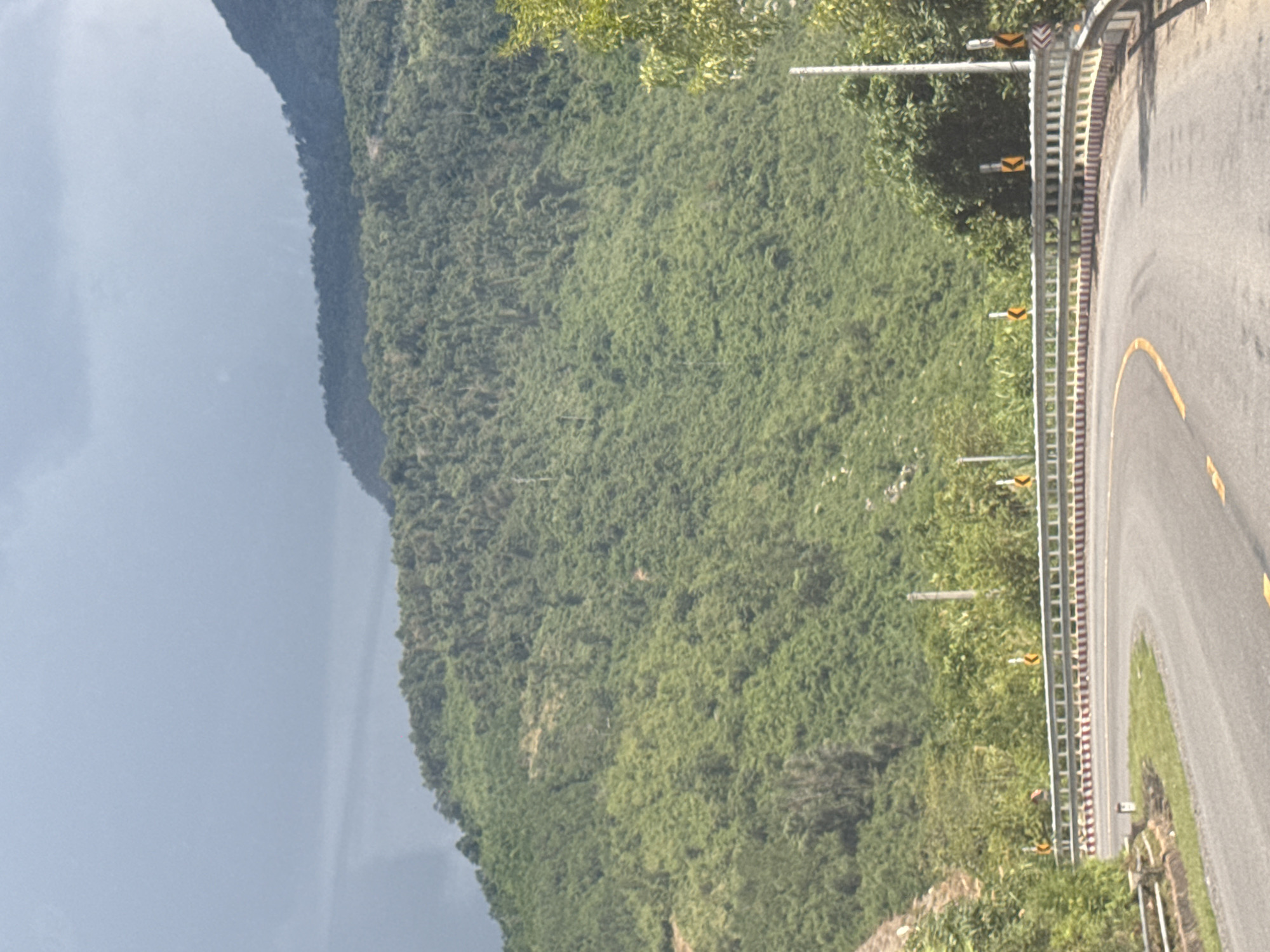 The image shows a winding asphalt road with a yellow line marking its center. The road curves to the left and appears to be on a mountain pass. A metal guardrail runs along the outer edge of the road, with warning signs indicating curves. The mountainside is densely covered with green vegetation, likely trees and bushes. The sky is overcast with dark clouds. The scene is likely in Nam Khanh Vinh, Vietnam, a region known for its mountainous terrain and winding roads.