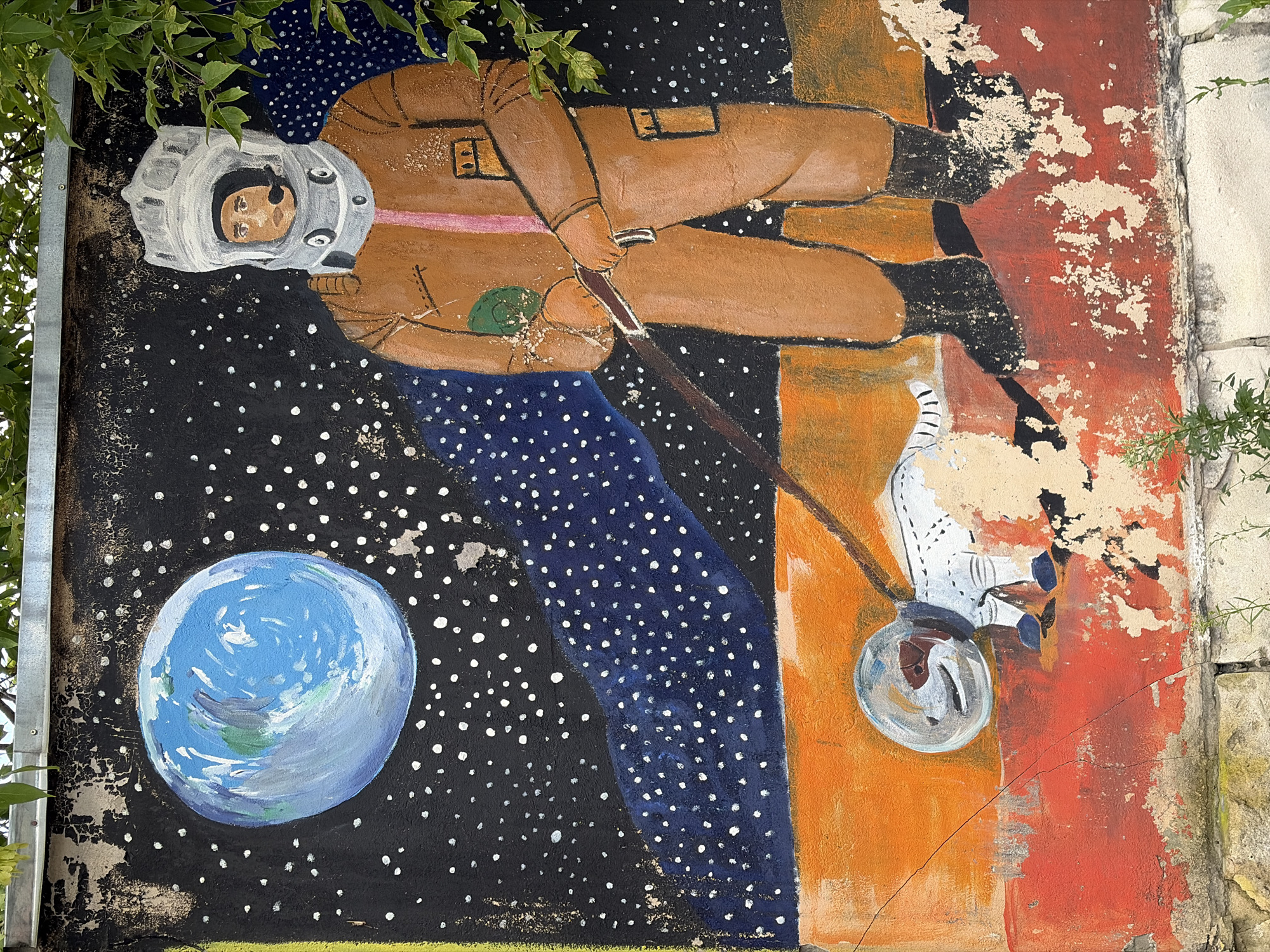 This is a detailed description of a mural located on a rough, weathered wall, likely in an outdoor urban environment, possibly in Moscow, Russia, given the context. The mural depicts a stylized scene of an astronaut walking a dog in space.
The mural's background is a dark, starry night sky, punctuated by numerous white dots representing stars. A large, blue and green representation of Earth is visible in the upper left quadrant. Below this, a swath of deep blue is painted, also speckled with white dots, suggesting a deeper part of space or a nebula.
The main subjects are a human astronaut and a dog. The astronaut is depicted in a bulky, orange spacesuit with a large, white helmet. The helmet has a clear visor through which a person's face, with brown hair and wearing a headset, is visible. The astronaut's suit features a pink stripe down the front and two yellow rectangular patches on the chest and leg. The astronaut is holding a leash attached to the dog.
The dog is painted in white with some brown markings around its face and paws. It is also wearing a spacesuit, complete with a clear bubble helmet and blue boots. The dog appears to be in a walking or slightly trotting pose, looking down. The leash connects the astronaut's gloved hand to the dog's helmet.
The foreground of the mural is a rough, textured landscape rendered in bold colors. The lower portion is a bright red, with sections of the plaster visibly chipped and peeling away, revealing lighter-colored concrete or stucco underneath. Above the red is a band of vibrant orange, with some cracks visible in the paint and underlying wall.
The mural itself shows signs of wear and tear. The paint is faded and chipped in many areas, particularly along the bottom edge and on the upper parts of the wall, where the black background is flaking. Overhanging the top of the mural are some green leaves from a tree or shrub, suggesting it's an outdoor location with vegetation nearby. The base of the wall appears to be constructed of rough, light-colored stone blocks.