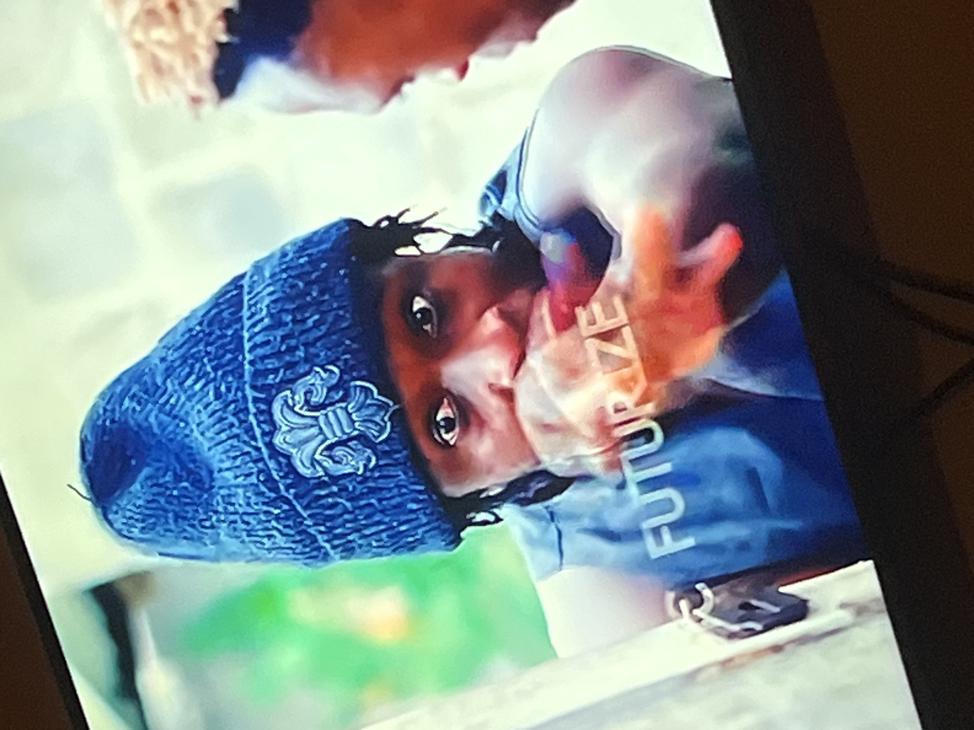 The image shows a close-up of two young men, one in the foreground and one in the background. The man in the foreground is wearing a blue knitted beanie with a fleur-de-lis emblem and a blue sleeveless shirt with the word "FUTURIZE" printed in white. He has dreadlocks and is looking directly at the camera with his left hand covering his mouth. The man in the background is partially visible on the right side of the frame, with light-colored hair and also looking towards the foreground. The background appears to be an outdoor setting with blurred green foliage and a light-colored textured wall. The image is a still from a video or film, possibly a scene from Gura Top, Nigeria.