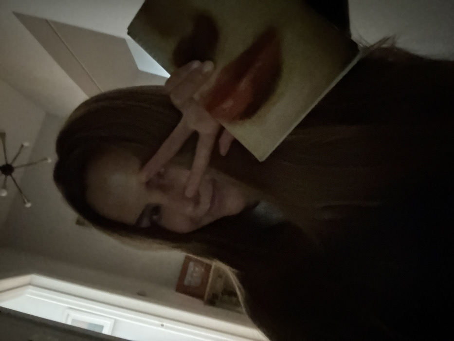 An indoor, dimly lit, close-up image features a female subject with long, dark brown hair, smiling and performing a peace sign (V-gesture) with her right hand, partially obscuring her face. Her left hand holds a light-colored rectangular object, possibly a book or small canvas, which displays a reddish-brown, organic shape resembling lips. The background, largely out of focus, reveals a residential interior. Visible elements include a light-colored doorframe or opening on the left, a piece of dark wooden furniture with a framed item on top, and a multi-armed modern light fixture with exposed bulbs mounted on the ceiling. No specific visual cues within the image indicate a precise location within Mexico City, Mexico.