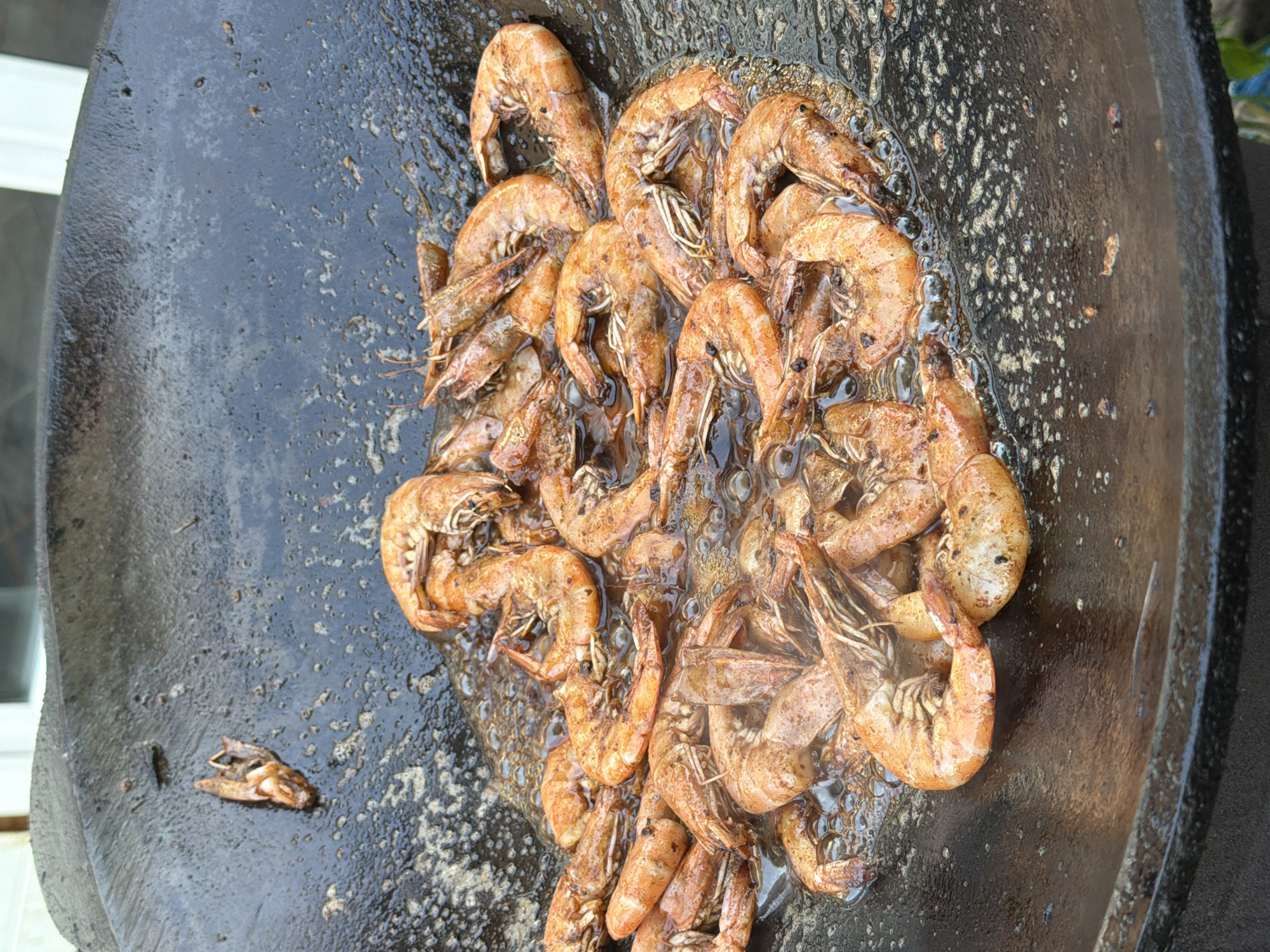 A close-up shot shows a large quantity of cooked shrimp sizzling in a black, round, cast-iron pan. The shrimp are reddish-orange and piled together in the center of the pan, with some appearing to be curled. They are immersed in a bubbling liquid, likely oil or sauce, which has created a textured surface with small bubbles and speckles. The pan itself has a worn, slightly textured appearance. In the upper left corner of the pan, a single, isolated shrimp is visible, separate from the main cluster. The background is blurred, but suggests an outdoor setting, possibly with a light-colored structure visible. This scene is likely from a food preparation area, possibly a street food vendor or outdoor kitchen. The location in Orenburg, Russia is not discernible from the visual information provided in the image.