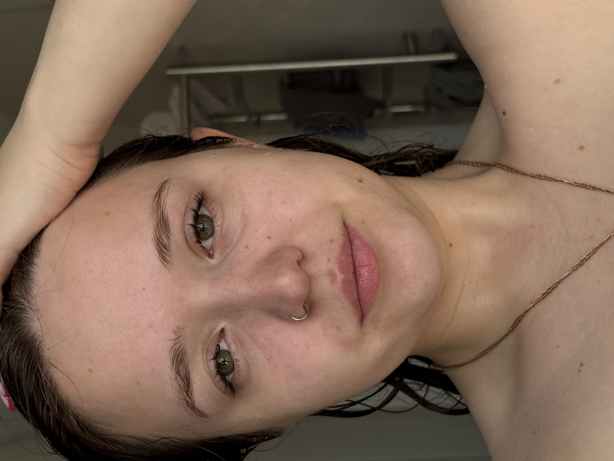 A close-up portrait of a woman with light skin, green eyes, and brown hair. She has a nose ring and is wearing a gold chain necklace. One hand is raised towards her head. The background is blurry, but includes what appears to be a towel rack. It is impossible to determine the location of the image as Izhevsk, Russia from the provided image alone.