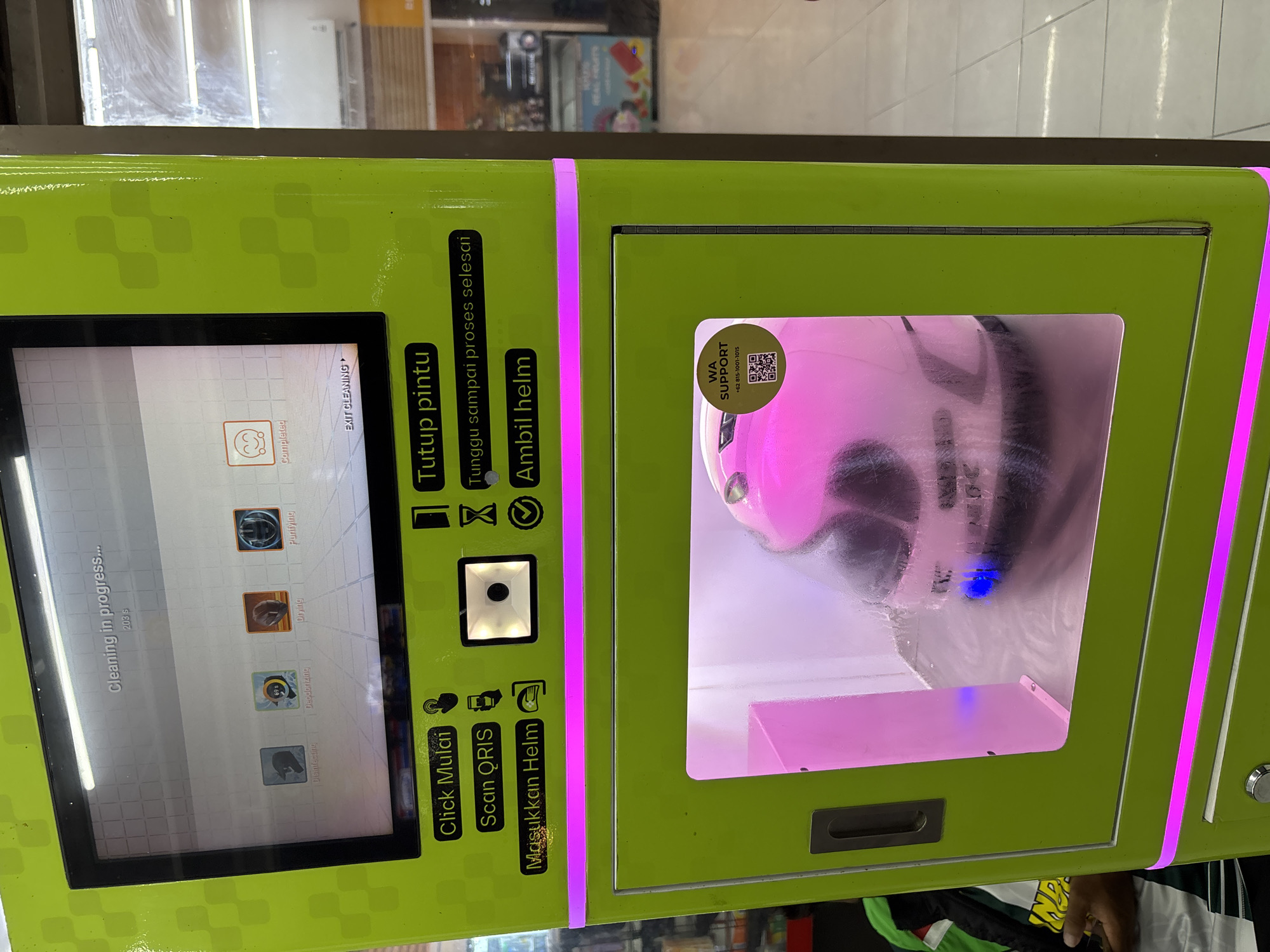 The image shows a helmet cleaning and disinfecting machine, likely located in Kuta Selatan, Indonesia, based on the Indonesian text on the machine and the WA support number (+62). The machine is green with purple accents. A screen displays "Cleaning in progress..." along with icons for Disinfecting, Deodorizing, Drying, and Purifying. The text "Click Mulai, Scan QRIS, Masukkan Helm" is visible, indicating the steps for using the machine. Another sign reads "Tutup pintu, Tunggu sampai proses selesai, Ambil helm," instructing users to close the door, wait for the process to complete, and then take their helmet. The machine has a compartment with a helmet inside, illuminated by a pinkish light, likely indicating the cleaning or disinfecting process.