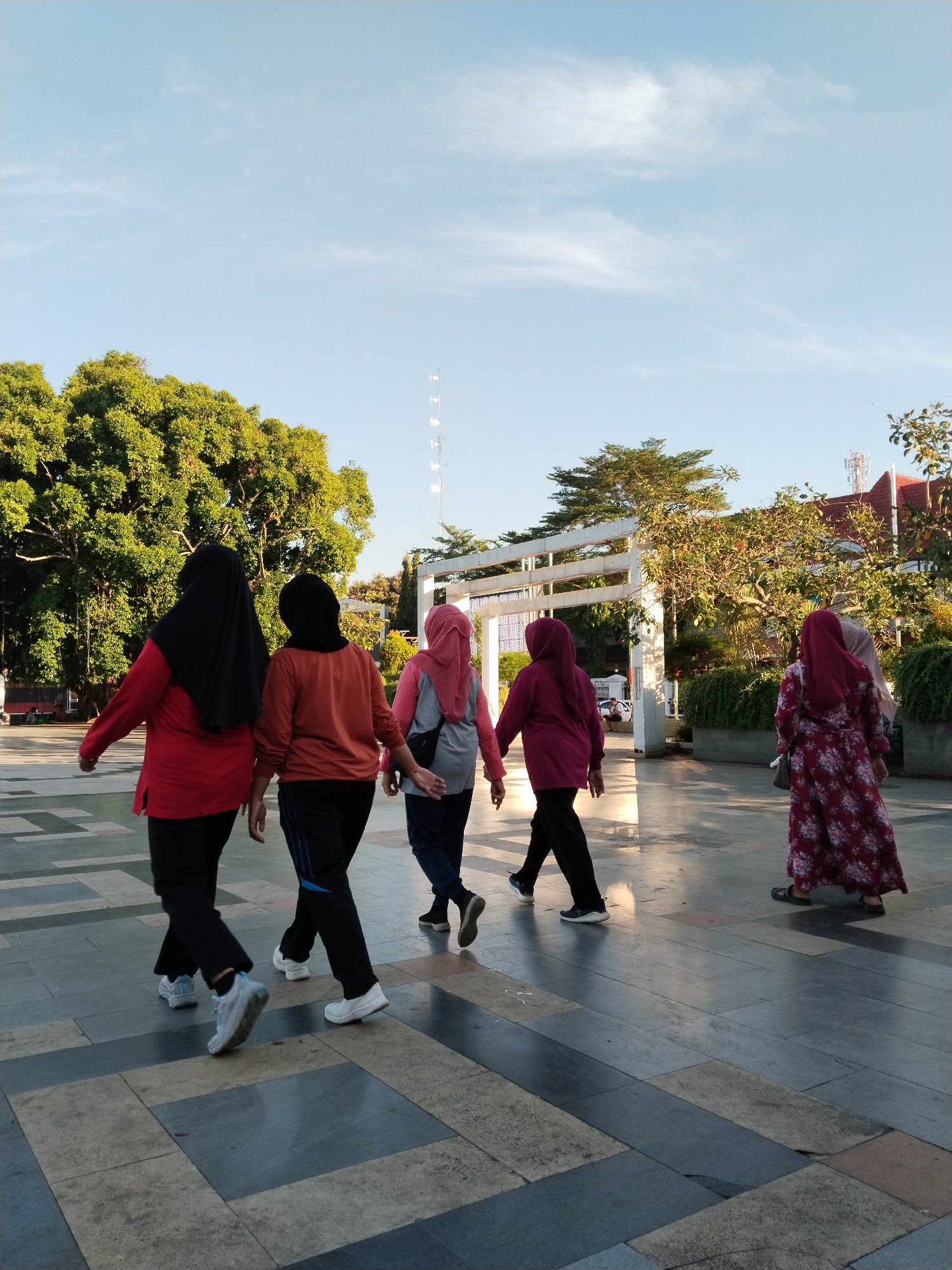 The image captures a group of five women walking outdoors in what appears to be a public space with a patterned paved ground. The women are dressed in modest clothing, with at least four of them wearing hijabs. The woman on the far left wears a red long-sleeved shirt and black pants. Next to her, a woman in an orange long-sleeved shirt and black pants walks. The third woman from the left wears a pink hijab and a gray top with a backpack. The fourth woman wears a maroon long-sleeved top and black pants. The woman on the far right is wearing a floral-patterned dress and a light-colored hijab. They are walking away from the viewer, towards a white archway structure. Behind the archway, trees and buildings are visible. A tall communication tower can be seen in the background. The sky is clear and blue with some scattered clouds. The scene suggests a casual walk or outdoor activity. The location is identified as Kecamatan Pati, Indonesia.