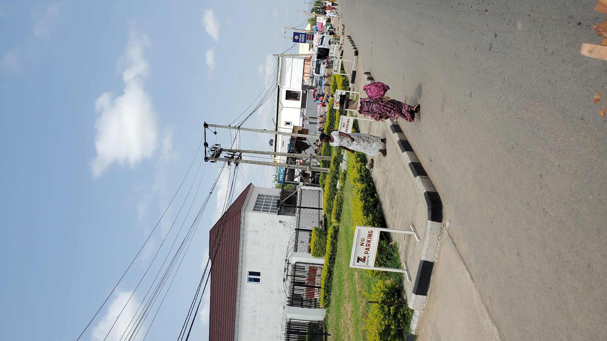 The image captures a street scene in Jalingo, Nigeria, under a bright, cloudy sky. On the left, a modern white building with a dark brown roof is visible, set behind a black metal fence and surrounded by neatly trimmed green bushes. Several power lines stretch across the upper portion of the image, supported by utility poles. A white sign with black lettering, reading "Z NO PARKING" with "ZENITH" in smaller text below "NO", stands prominently on the sidewalk.
Several people are present. Two women are walking on the sidewalk. One woman, wearing a light-colored, patterned dress, walks towards the right, carrying a pink bag over her shoulder. Another woman, in a light-colored, long dress, stands facing left on the sidewalk, near the "NO PARKING" sign. Further down the street, more people and vehicles are visible, including cars and vans. Another "NO PARKING" sign can be seen further down the road. The street itself is paved, with a curb that has alternating black and white painted sections. The environment appears to be a commercial or residential area with infrastructure typical of a Nigerian city.