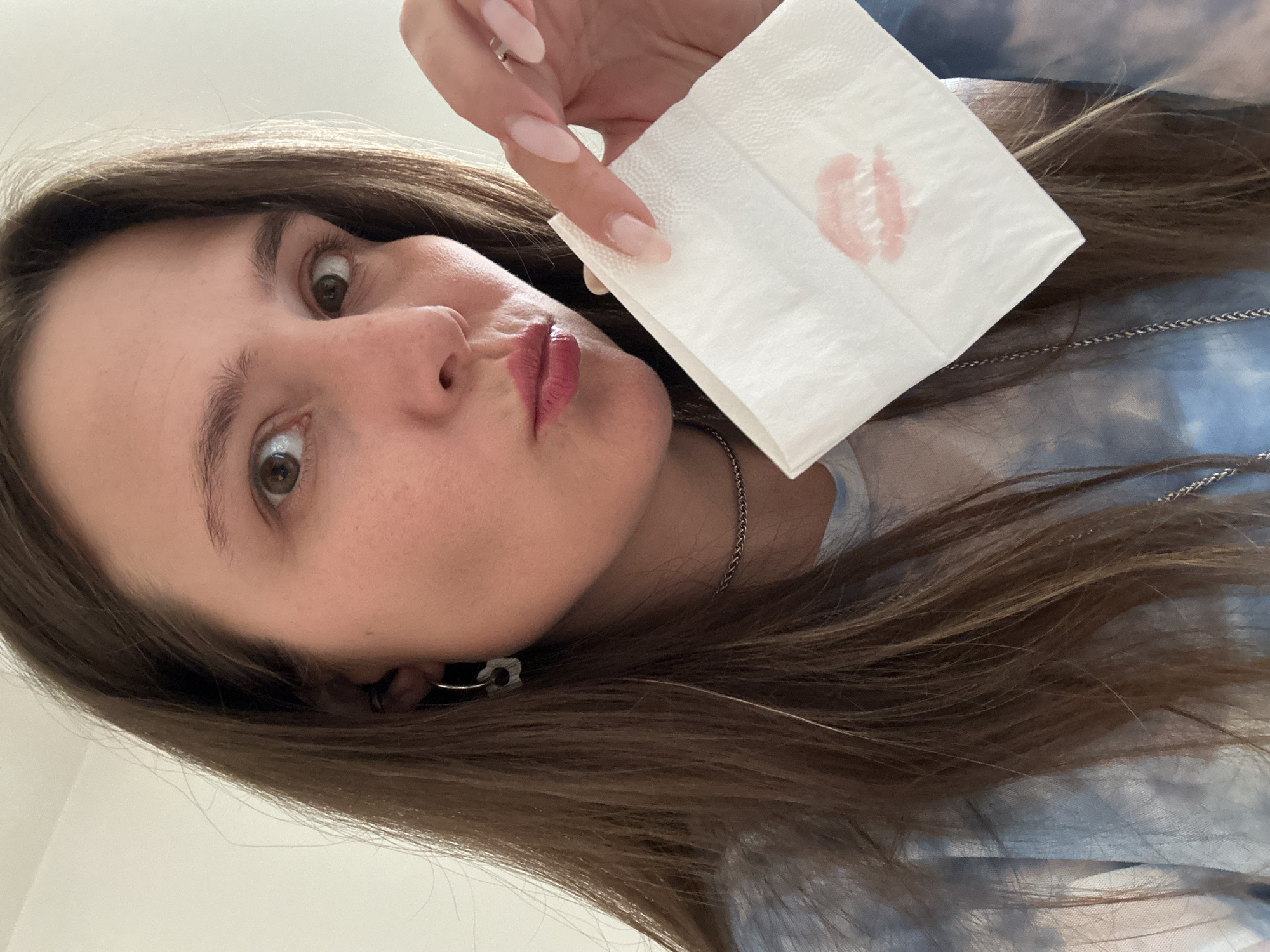 A woman is holding a white napkin with a lipstick print on it. The background shows the color of a light blue tie-dye shirt. The image appears to have been taken indoors. There is no evidence to suggest it was taken in the city of Šušanj, Montenegro.