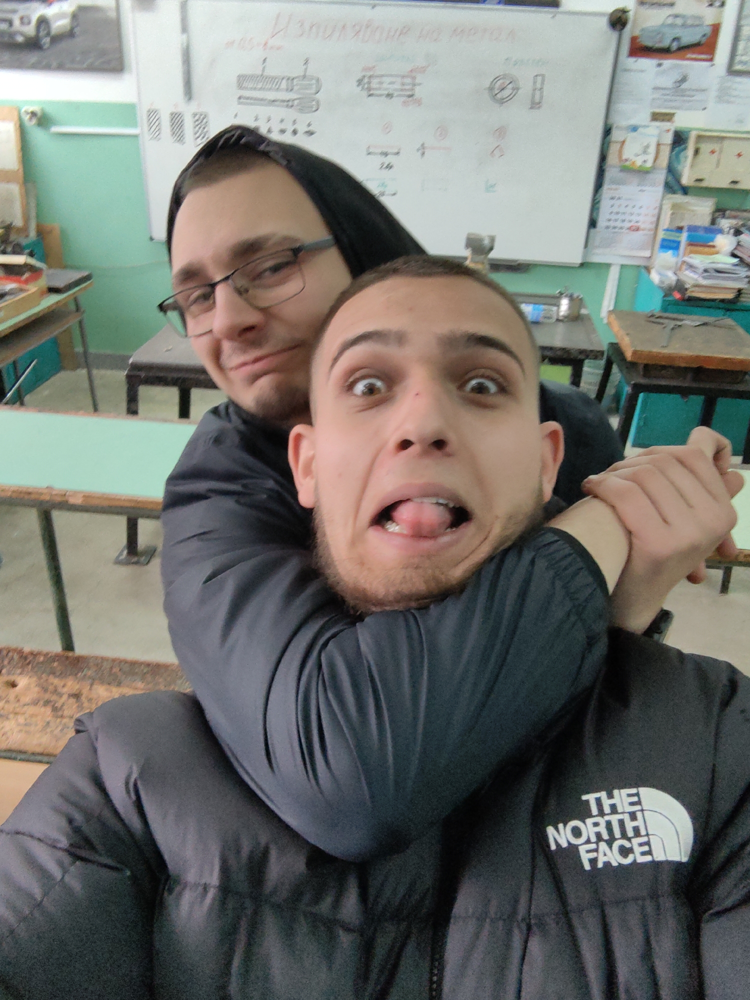 Two young men are posing for a selfie in what appears to be a classroom setting. The man in the foreground, wearing a black "The North Face" jacket, is making a funny face with his tongue sticking out and eyes wide open. He is being playfully choked from behind by another man wearing glasses and a black jacket with a hood. The classroom has green walls and visible desks. A whiteboard with technical drawings and text is in the background. The presence of Bulgarian text on the board suggests that the location is in Bulgaria, likely in Sofia given the request.