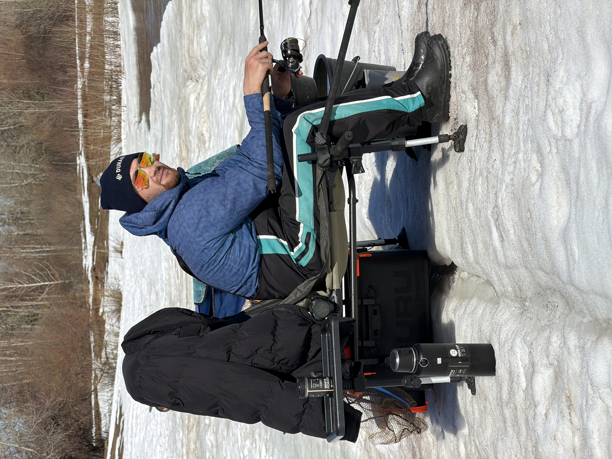 A man is seated on a fishing chair, holding a fishing rod, on a snow-covered area near a wooded area. He wears a blue patterned jacket, black pants with a light blue stripe, black boots, and a black knit cap with white lettering. He has orange-tinted sunglasses on. Fishing equipment surrounds him, including a black fishing box and a metal rod support. The wooded area in the background has bare trees. The image was taken in Noviy, Russia.